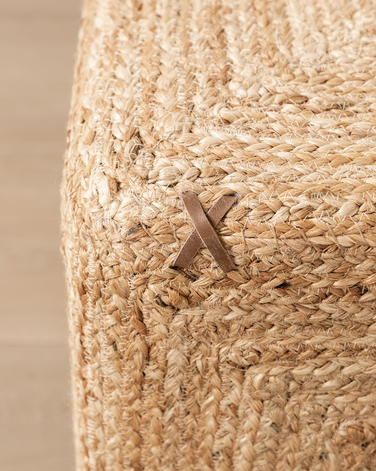 Close-up of the corner of the Kelvin Woven Ottoman, a hand-woven jute piece with a decorative tan leather X-shaped detail stitched on the surface.