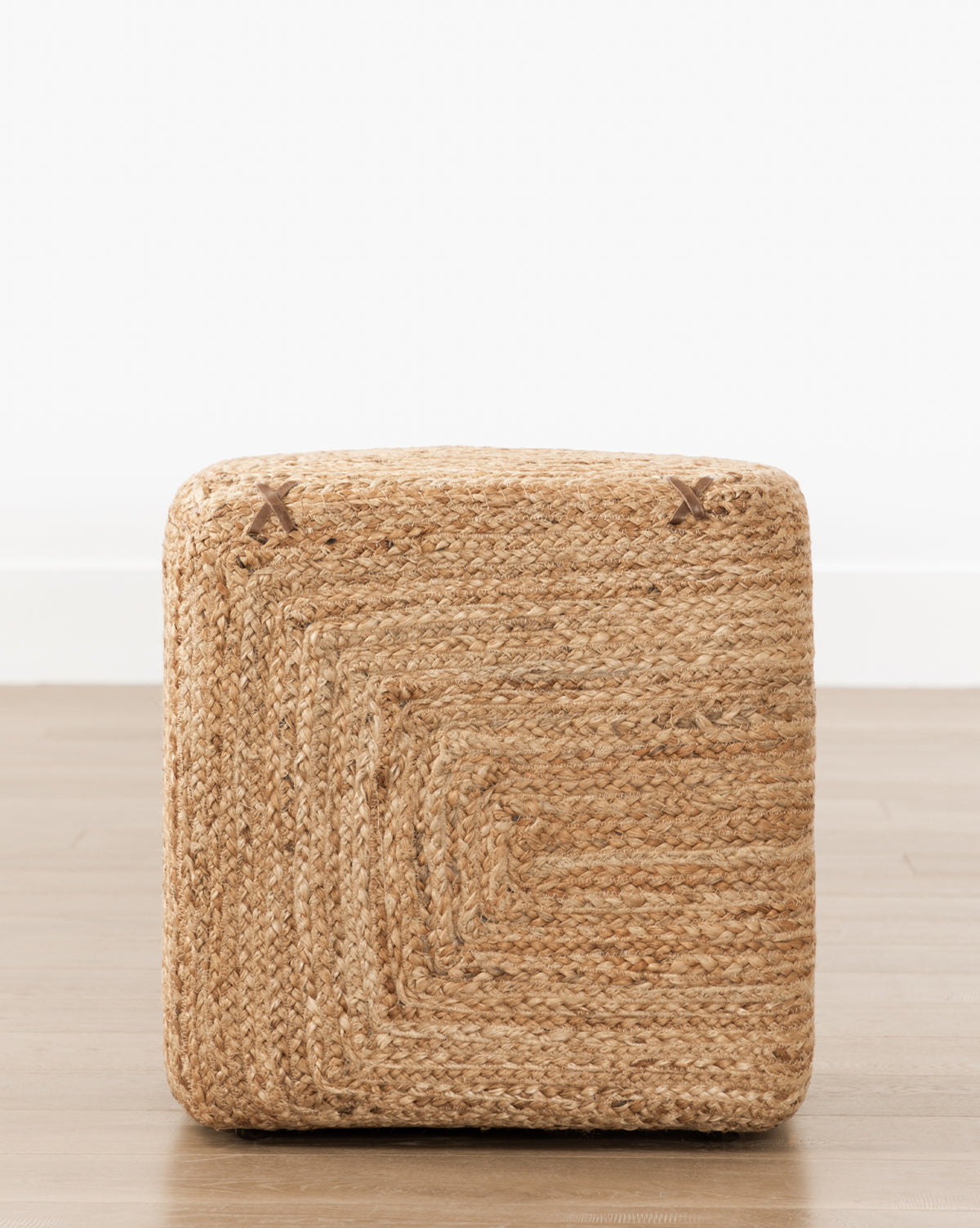 A hand-woven jute Kelvin Woven Ottoman sits on a light wooden floor by a plain white wall. Its natural fiber texture and subtle stitching details make this ottoman a versatile accent for any space.