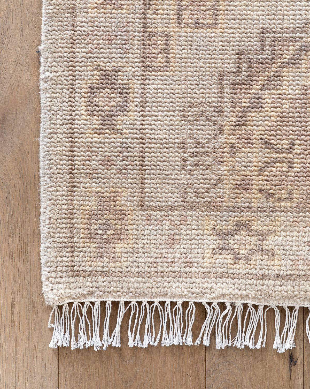 A close-up of the corner of the beige Kenna Hand-Knotted Wool Rug, featuring a subtle geometric pattern and fringed edges, shown on a light wood floor, by McGee & Co.