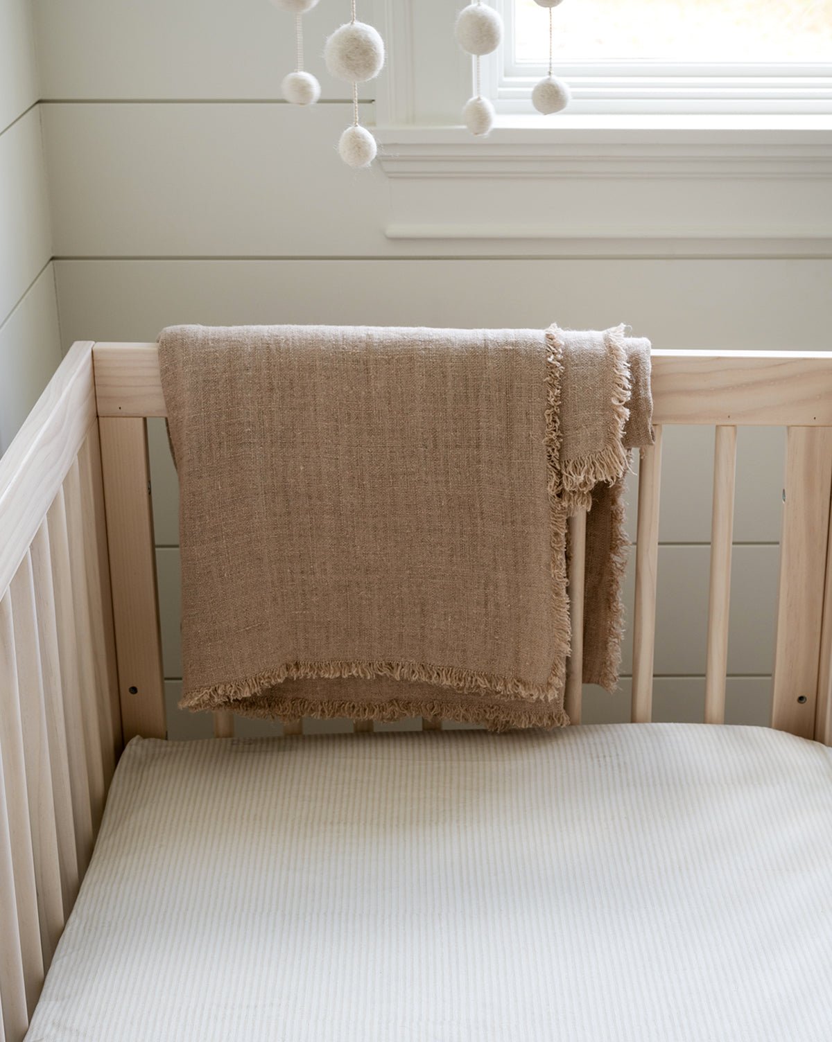 A beige textured blanket with fringes hangs over a light wooden crib dressed in an Easton Crib Sheet, cream-striped, below a window and a white pom-pom mobile, adding cozy charm to your baby’s room.