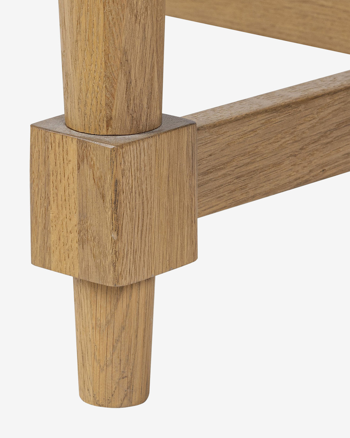 Close-up of a Kyler Oak Extension Dining Table leg with a square base and round, tapered lower section, joined to a horizontal support bar; all parts feature a light natural oak wood finish, by McGee & Co.