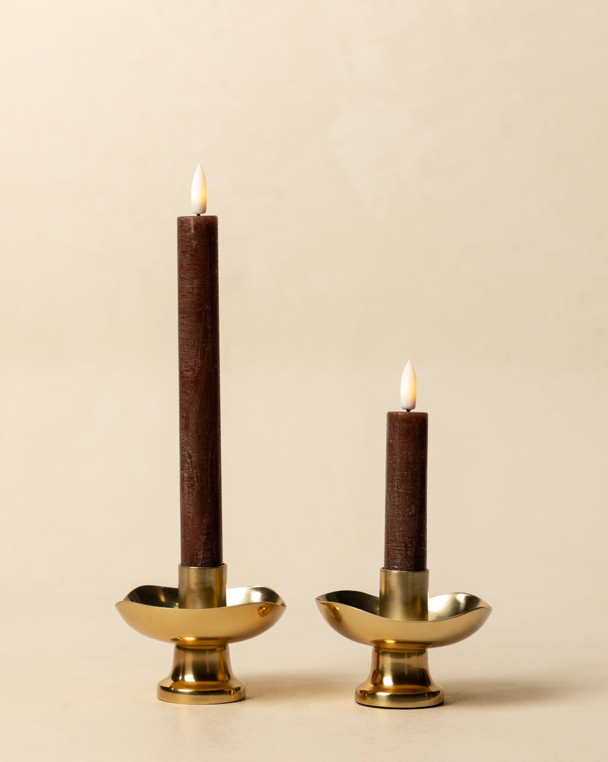 The LED Wax Taper Candles (Set of 2), in warm brown and varying heights, sit in gold holders on a beige background, offering the cozy glow of real tapers with safe, elegant LED flicker, by McGee & Co.