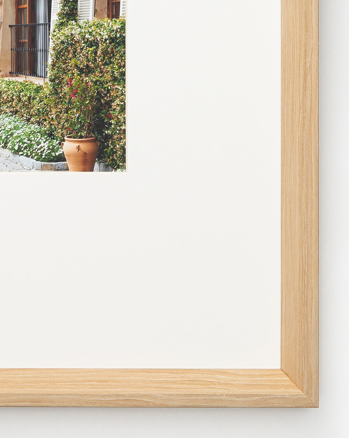 A close-up of a natural woodgrain frame with a white mat showcasing the giclee print La Residencia Deia, featuring a clay pot and greenery in an outdoor garden setting.