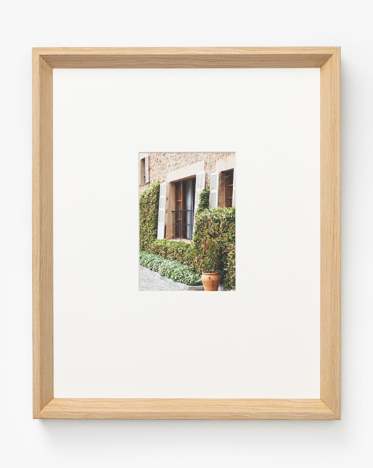 A framed photo titled La Residencia Deia shows a rustic building facade with a wooden door, shuttered window, green ivy, and terracotta pot. It’s presented in a wide white mat and natural woodgrain frame.