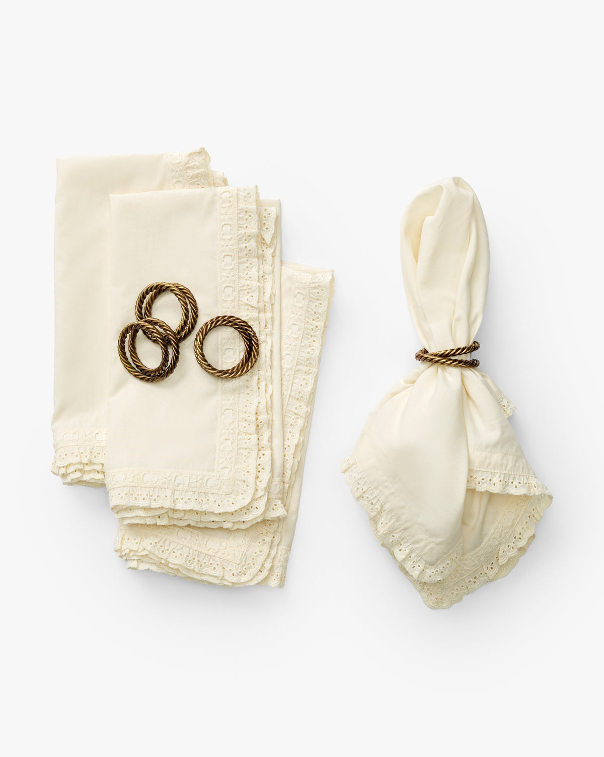 Four folded cream-colored Lace Trim Napkins (Set of 4) with delicate lace edges are stacked on the left, topped with three gold napkin rings. On the right, a single napkin is wrapped in a gold ring, all arranged on a white background, by McGee & Co.
