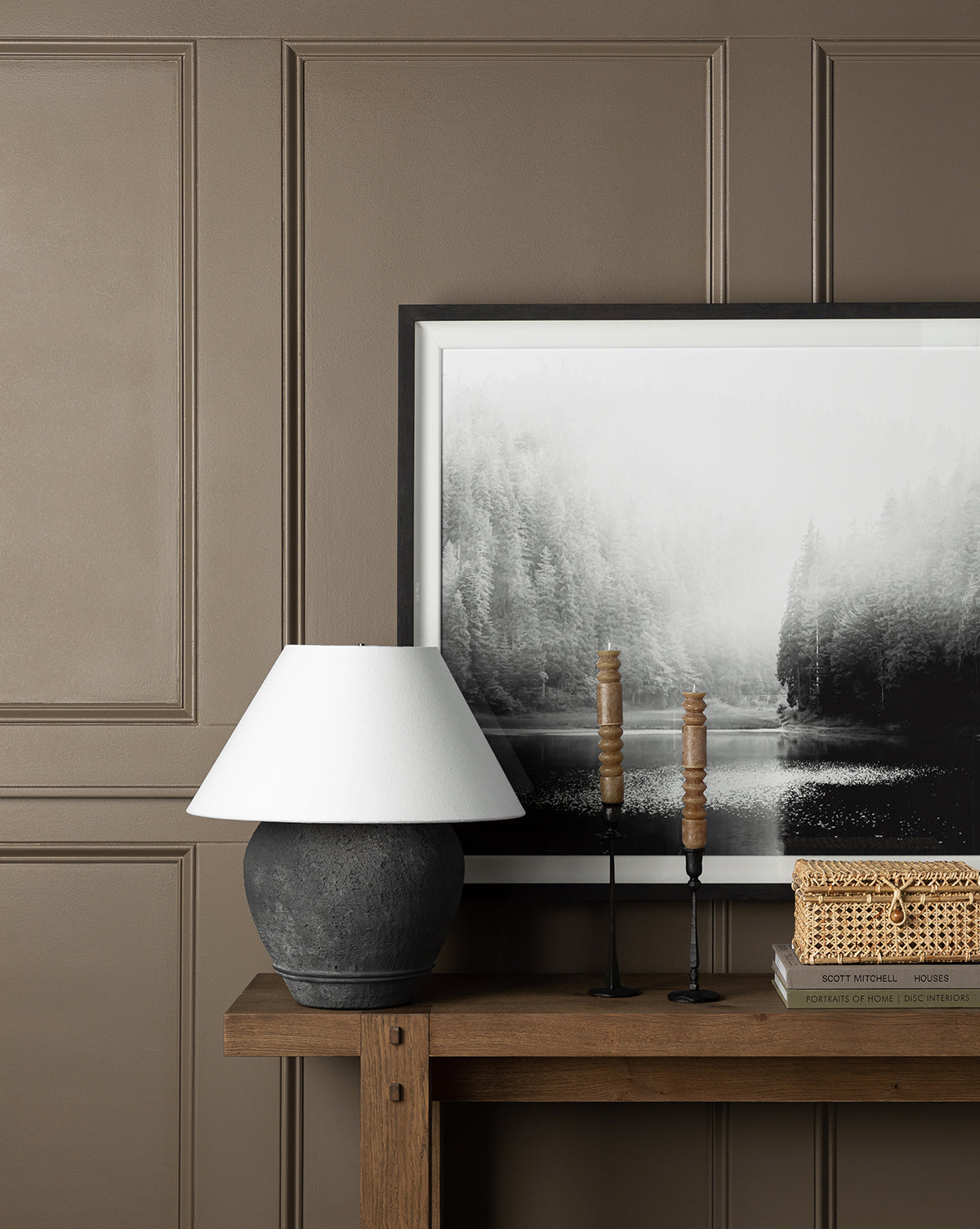 A console table displays a black lamp, two tall candlesticks, a woven box, and stacked books. Behind them hangs the Lakeside black-and-white wall art, depicting a misty forest and lake scene.
