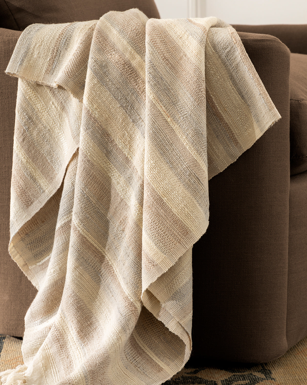 The Laney Stripe Throw by TAL HANDWOVEN CLASSICS LLP, in beige and cream stripes with tassels, is draped over a brown armchair, adding textured luxury and warmth to your space. McGee & Co.