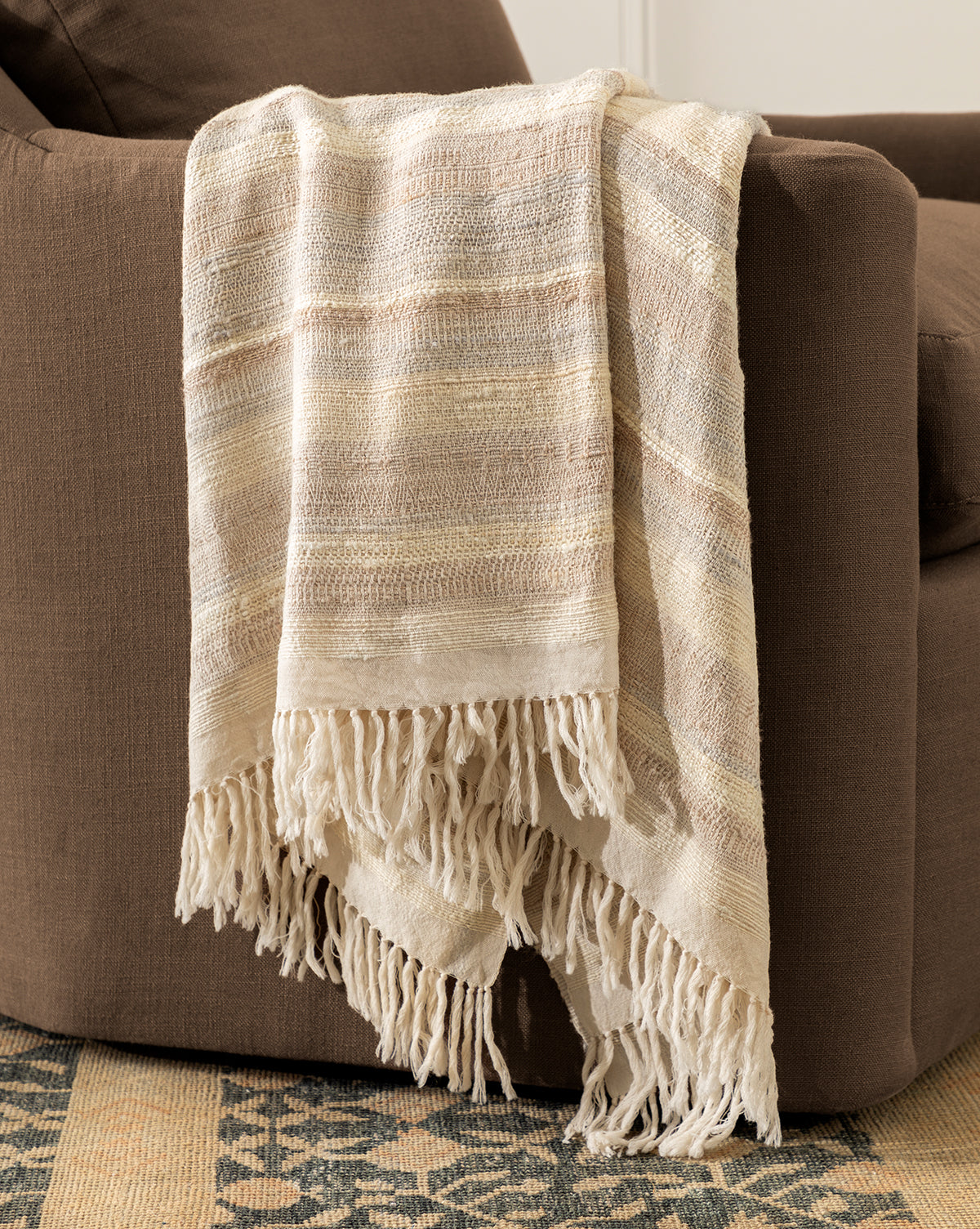 The Laney Stripe Throw by TAL HANDWOVEN CLASSICS LLP, featuring cream and beige stripes with fringed edges, is draped over a brown upholstered chair, with a patterned rug partly visible on the floor. McGee & Co.