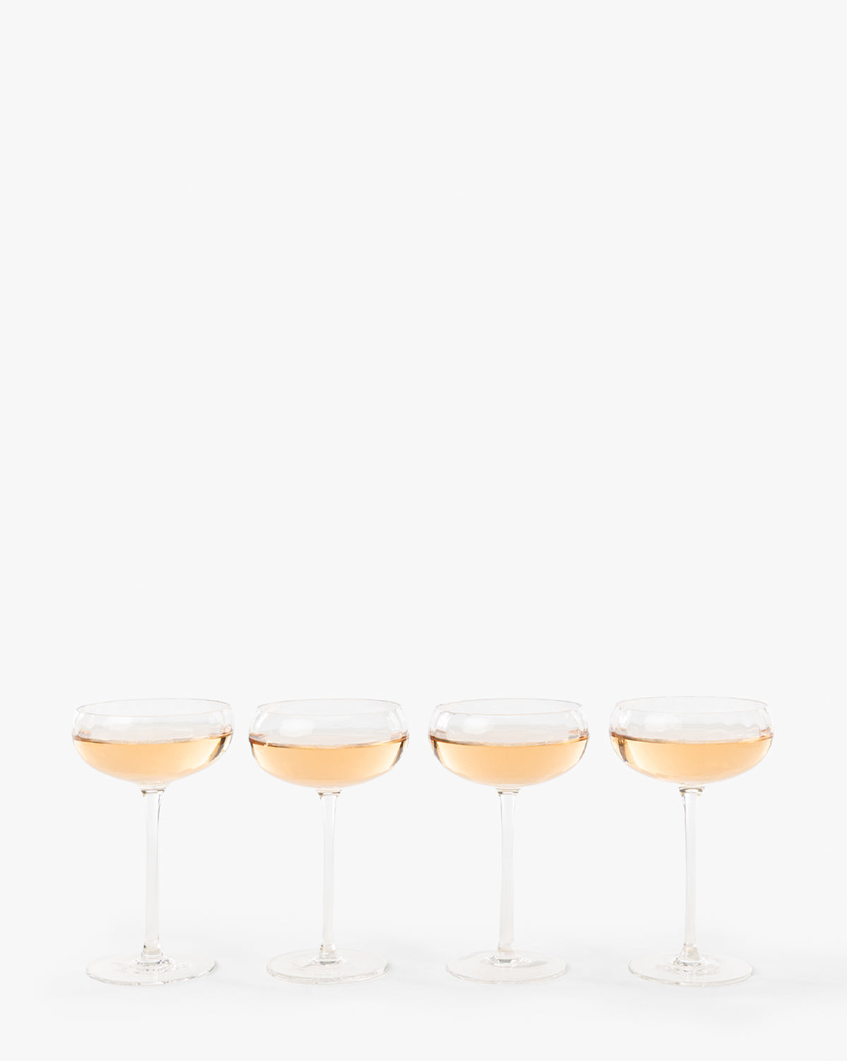 A set of four Langford Coupes, filled with a light pink sparkling beverage, are lined up against a plain white background, highlighting the elegance of this glassware set—perfect for any celebration, by McGee & Co.