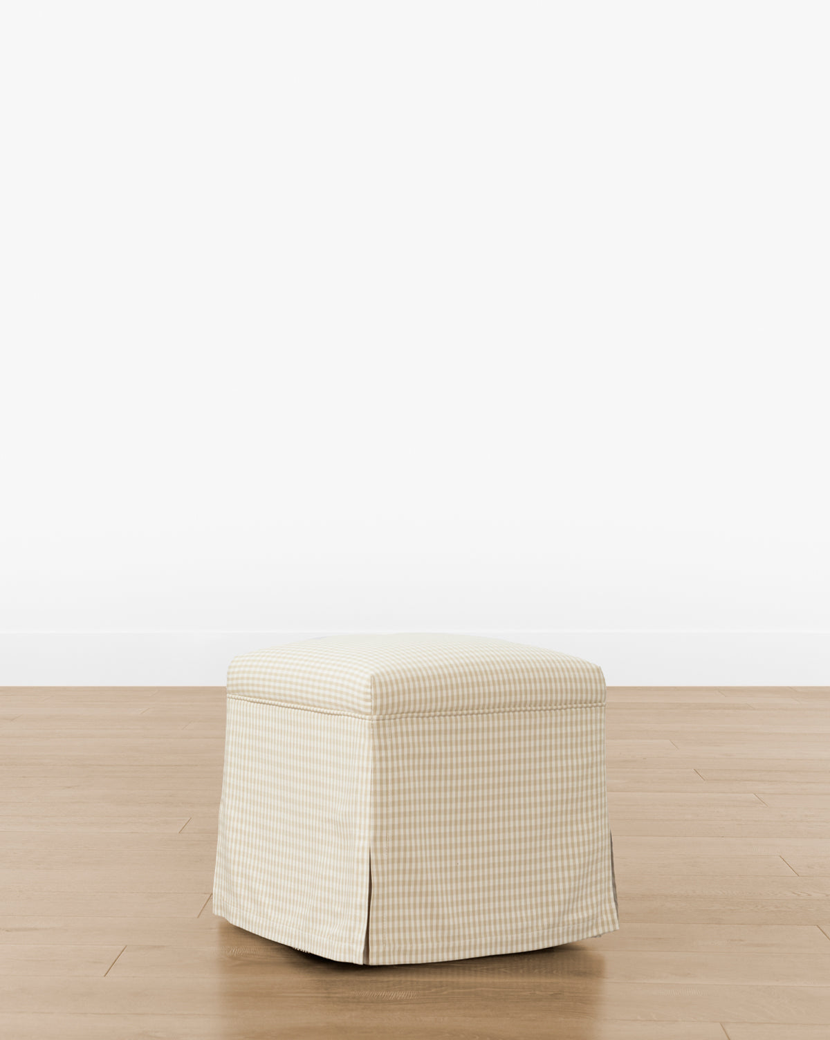 The Makerspalm Latimer Ottoman (Ready to Ship) features a beige and white gingham-patterned slipcover with pleated corners, shown on a light wood floor against a plain white background. McGee & Co.