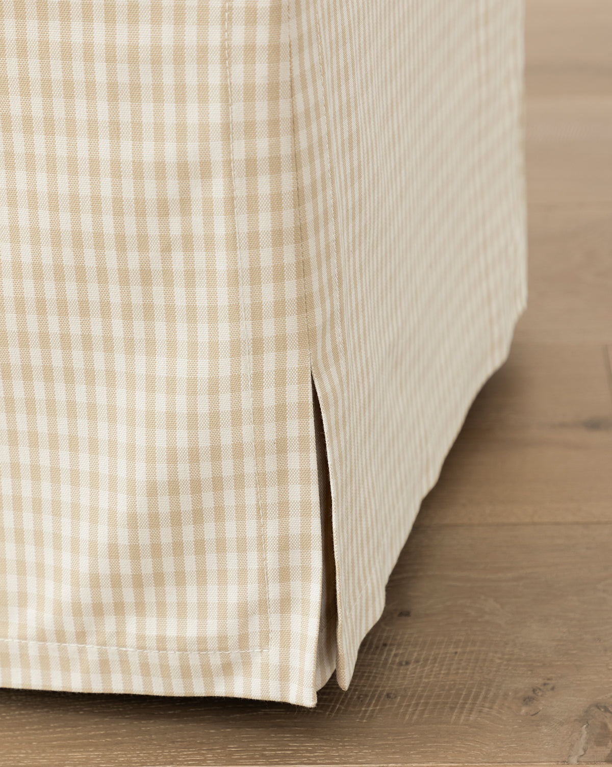 Close-up of the Makerspalm Latimer Ottoman (Ready to Ship), featuring beige and white gingham-patterned fabric with a tailored corner and vertical slit, elegantly placed on a light wooden floor. McGee & Co.