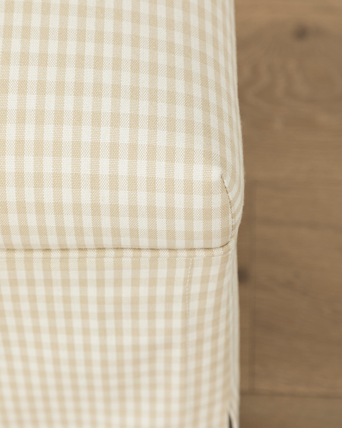 Close-up of the Makerspalm Latimer Ottoman (Ready to Ship), featuring beige and white checkered upholstery, showcases its quality craftsmanship as a stylish accent piece, set against a wooden floor. McGee & Co.
