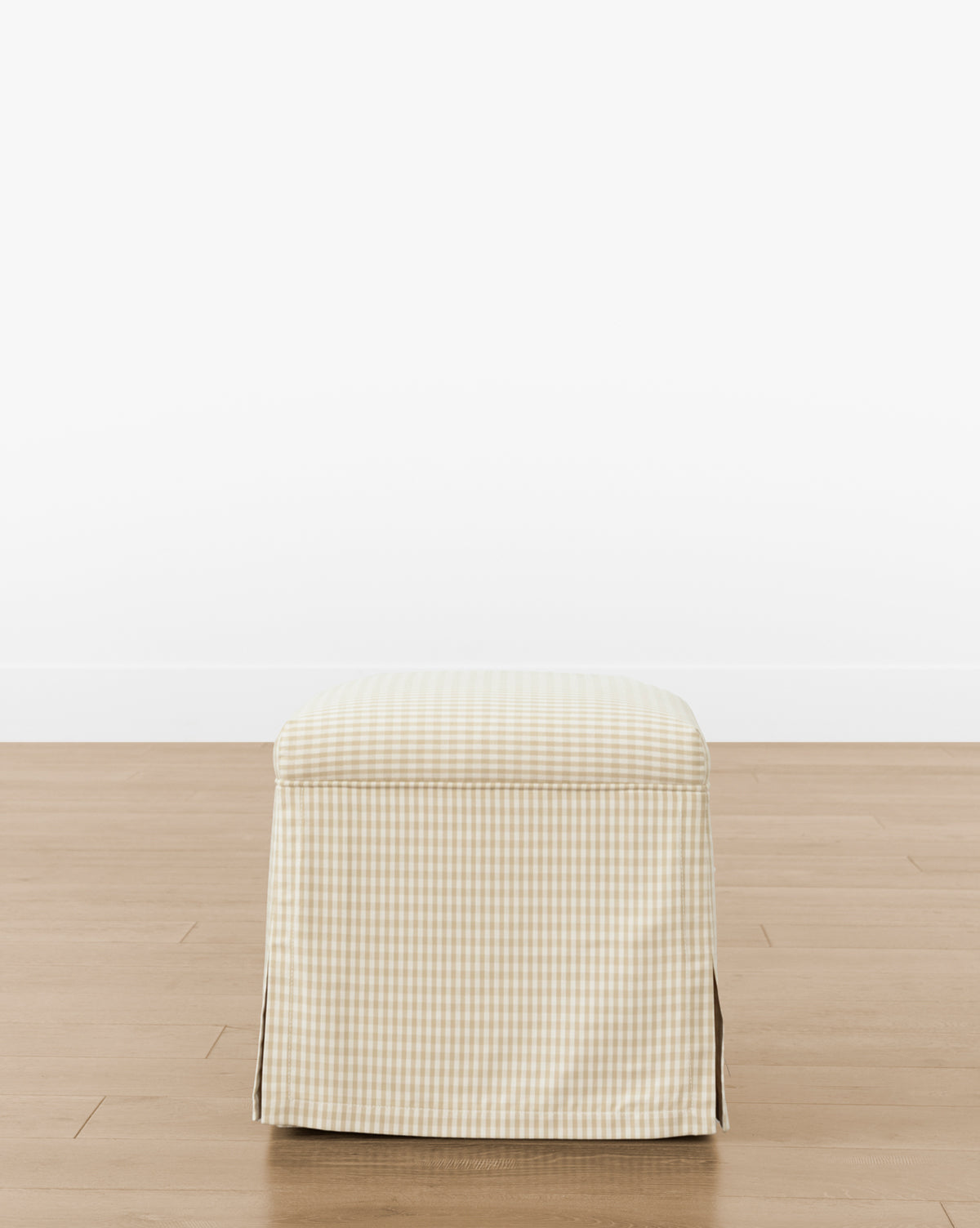 The Makerspalm Latimer Ottoman (Ready to Ship) features high-quality beige and white checkered fabric with a skirted cover, adding functional style to any room. Shown on a wooden floor against a white wall. McGee & Co.