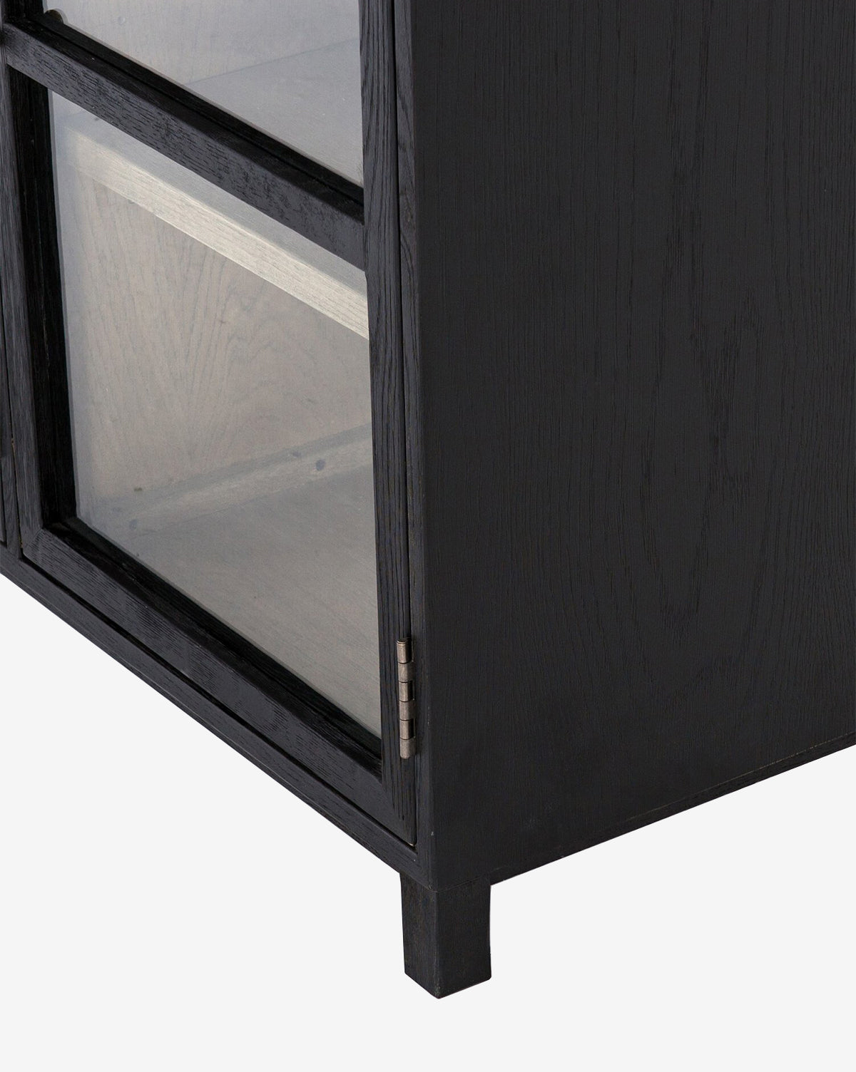 Close-up of the bottom corner of the Laura Sideboard in black oak, featuring glass panel doors, a visible door hinge, and part of the interior shelf.
