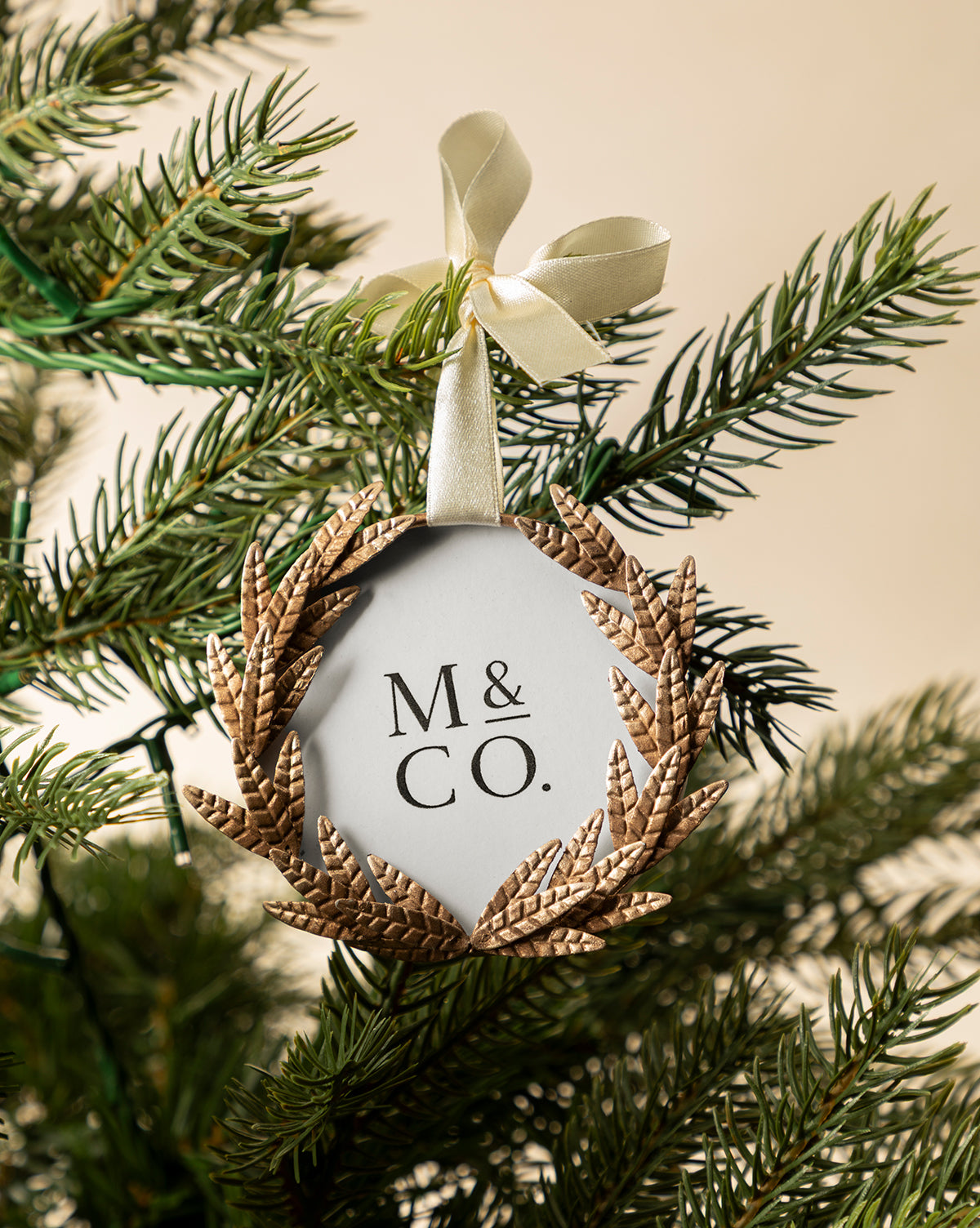 The Laurel Picture Frame Ornament features "M & CO." in black, framed by a gold laurel wreath and hung with a cream ribbon bow—an elegant, sentimental holiday gift that adds charm to any green Christmas tree branch, by McGee & Co.