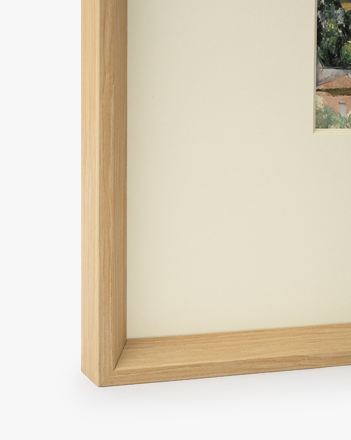 Close-up of a museum-quality natural woodgrain frame with cream matting, showing a small section of Le Village artwork in the upper right corner on a white background, by McGee & Co.