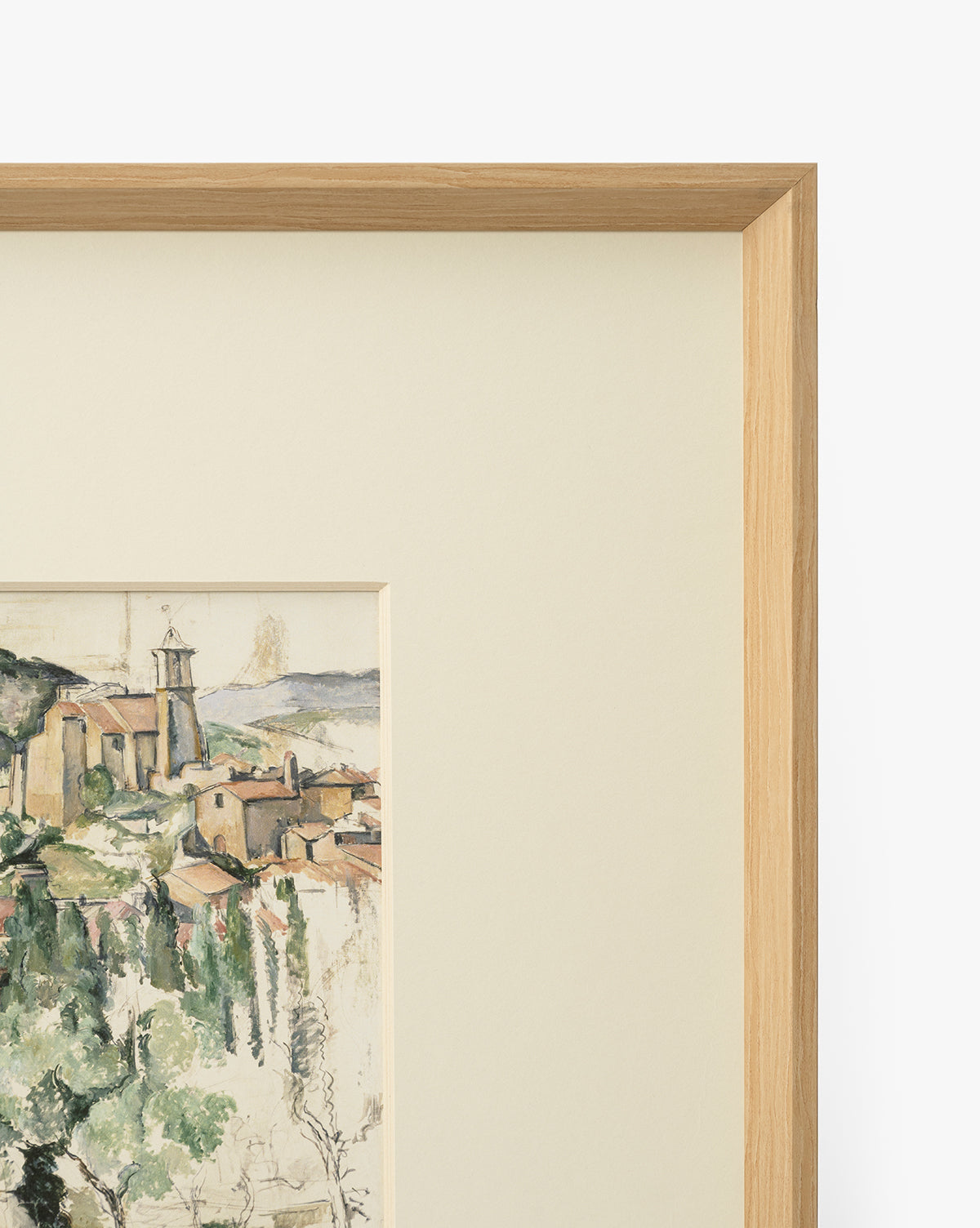 A close-up of Le Village, a museum-quality giclee print, shows part of a hillside village with buildings and greenery, accented by a light mat and an elegant natural woodgrain frame, by McGee & Co.