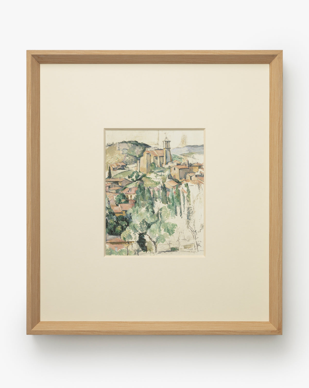 Le Village is a framed watercolor giclee print featuring a hillside town with clustered buildings, church steeple, and soft greenery. Museum-quality, its centered in a wide off-white mat and set in a natural woodgrain frame, by McGee & Co.