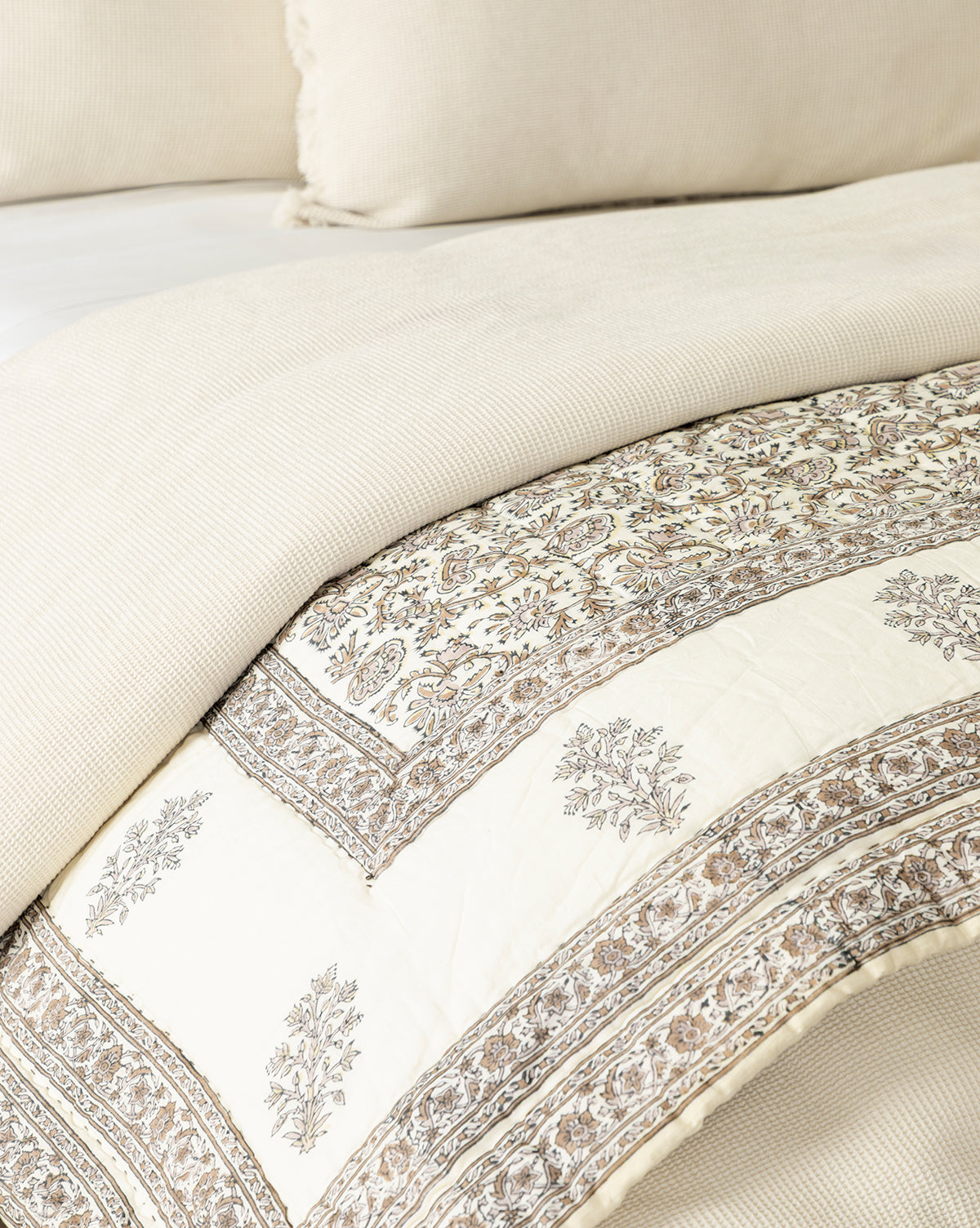 A close-up of a bed with cream-colored pillows and a textured beige duvet, partially folded to reveal the Leander Quilt and a cream and brown floral-patterned blanket underneath, adding charm to the bedding.