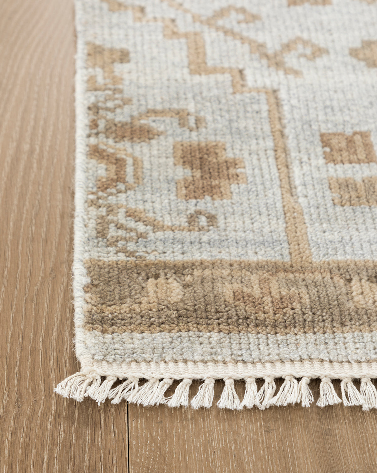 A close-up of the Lexington Hand-Knotted Wool Rug shows its geometric beige and light brown pattern on a wooden floor, finished with short white fringe along the edge, by McGee & Co.