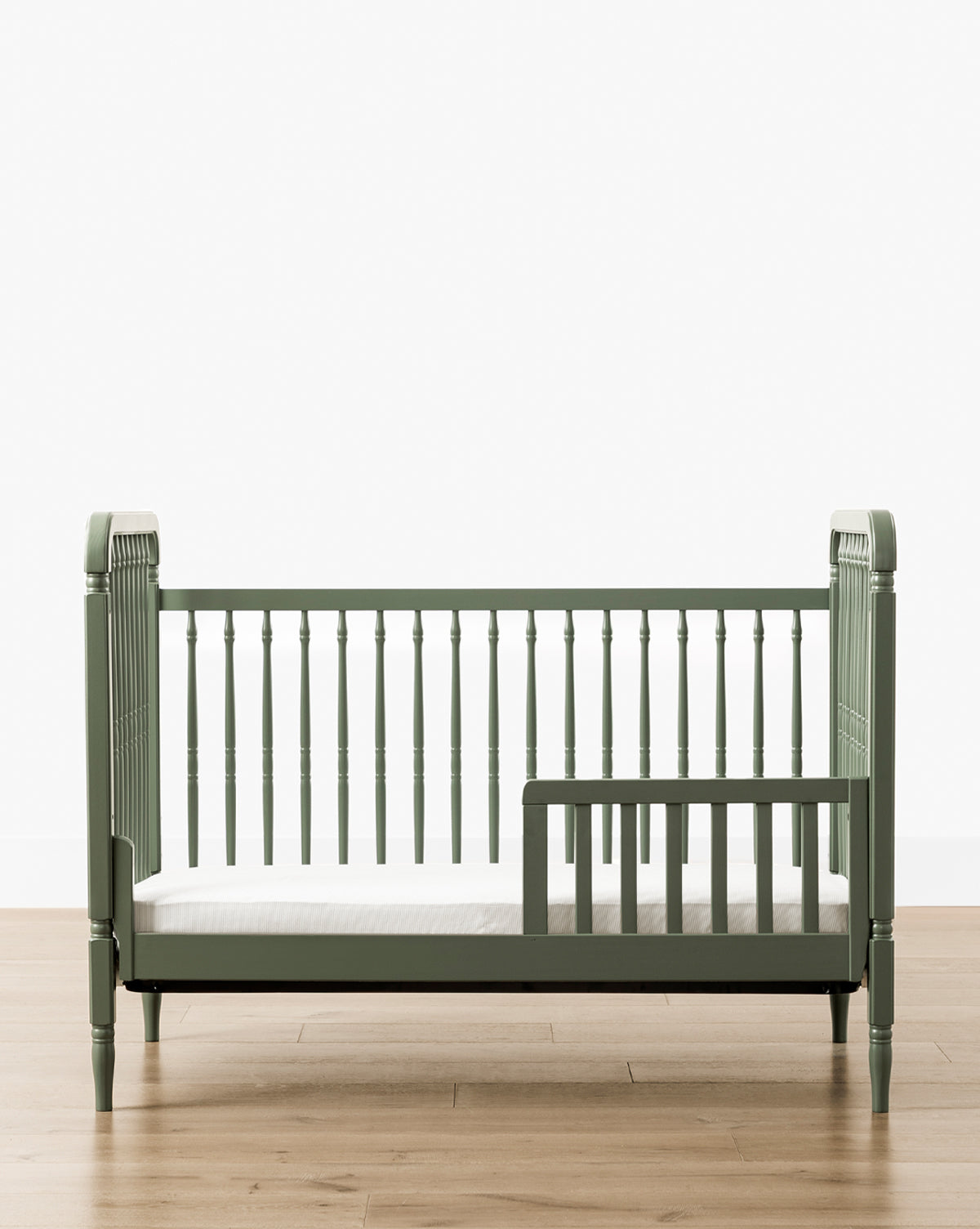 Liberty 3-in-1 Convertible Spindle Crib with Toddler Bed Conversion Kit in green, crafted from solid pine with vertical slats, low safety rail, and white mattress, shown on a light wood floor against a plain white background by McGee & Co.