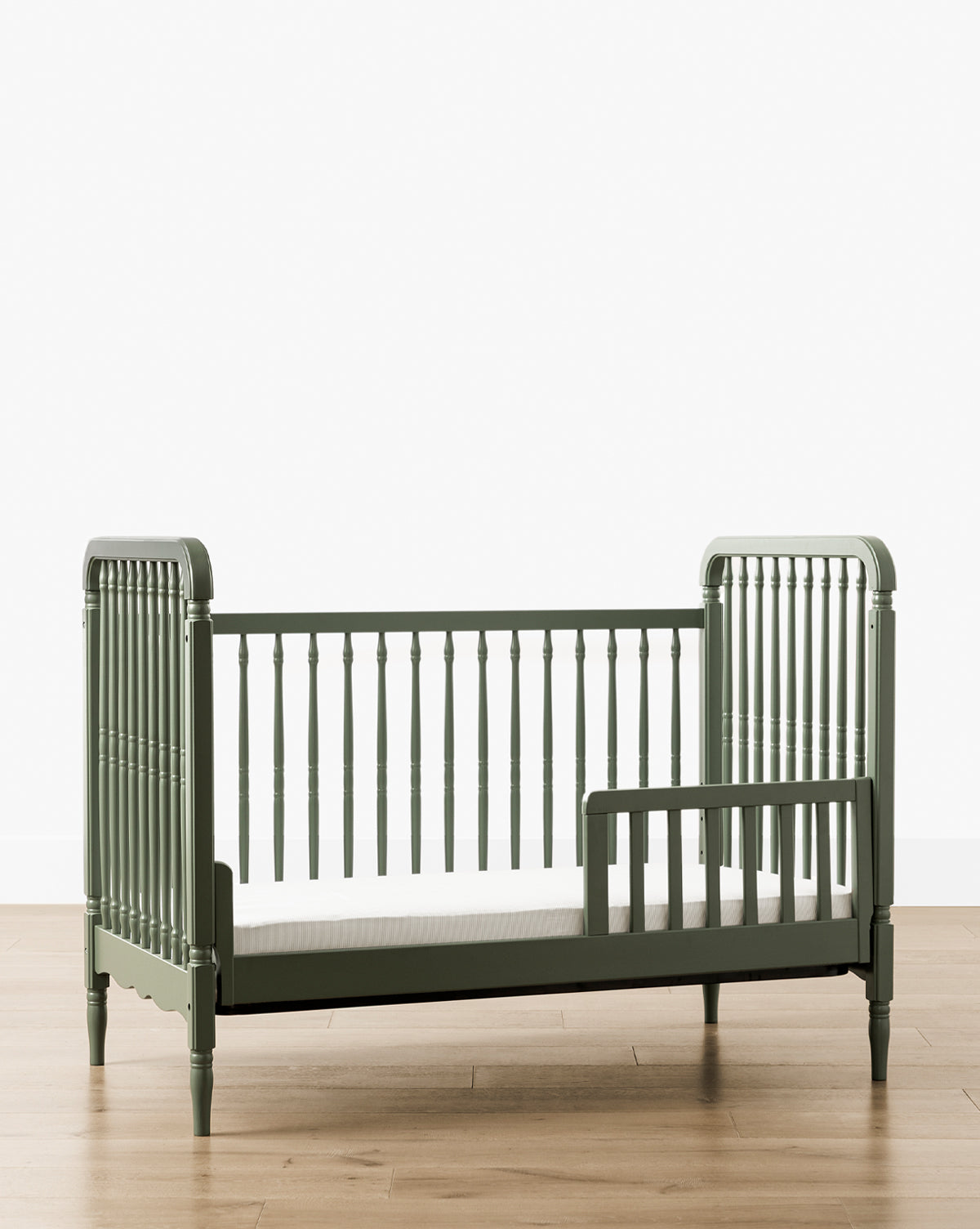 The Liberty 3-in-1 Convertible Spindle Crib with Toddler Bed Conversion Kit, crafted from solid pine and featuring slatted sides plus a partial safety rail, is shown on a wooden floor against a white background, by McGee & Co.