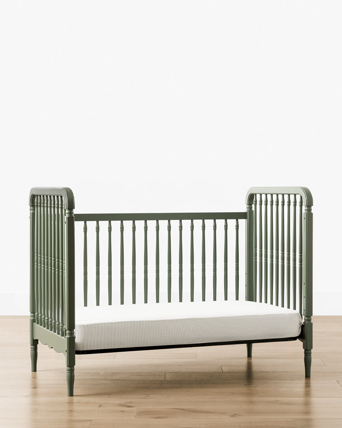 The Liberty 3-in-1 Convertible Spindle Crib with Toddler Bed Conversion Kit, shown in green solid pine with slatted sides and a white mattress, stands on a light wood floor against a plain white background by McGee & Co.