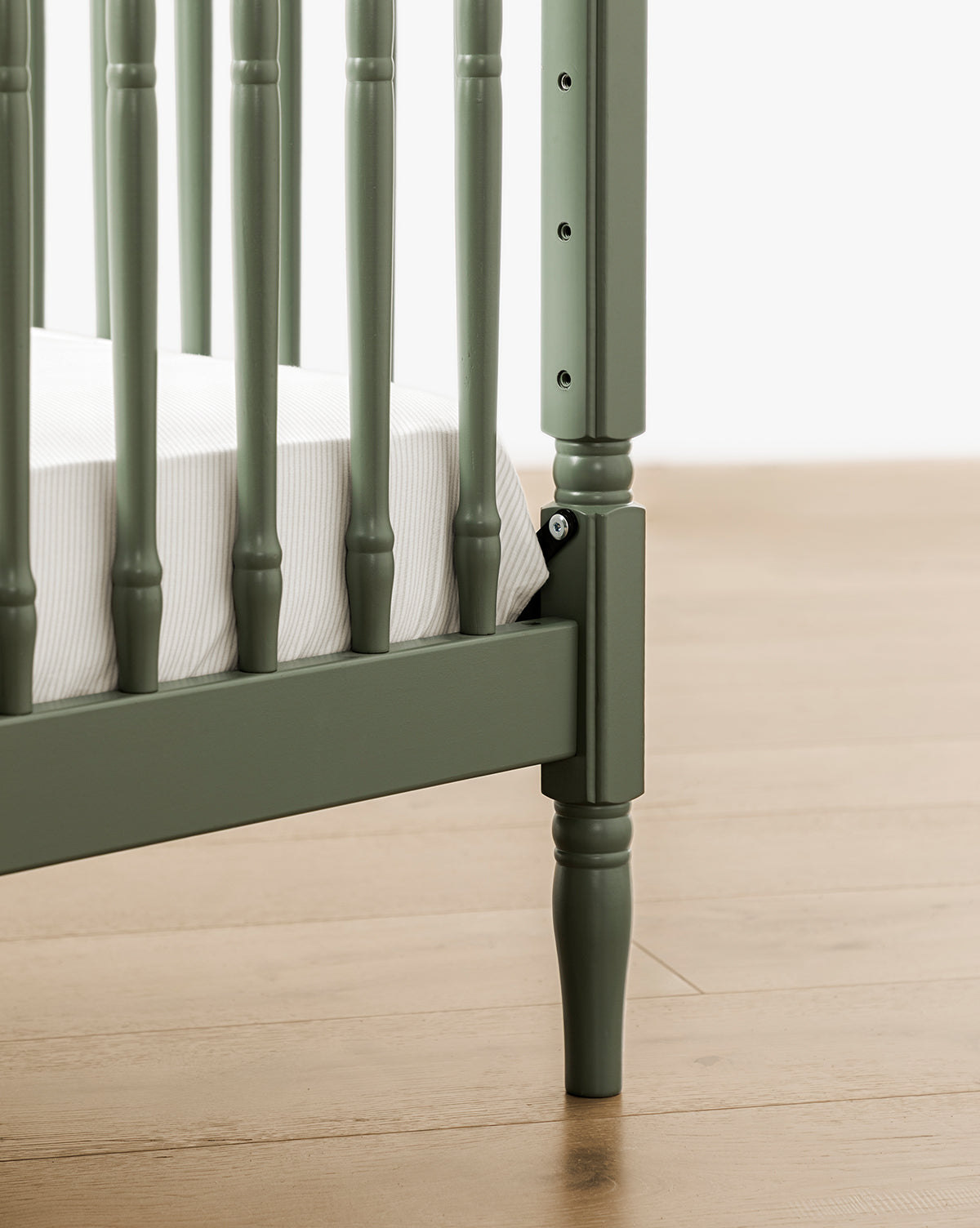 Close-up of the Liberty 3-in-1 Convertible Spindle Crib with Toddler Bed Conversion Kit in green solid pine, featuring vertical slats, a white mattress, and light wood flooring in the background, by McGee & Co.