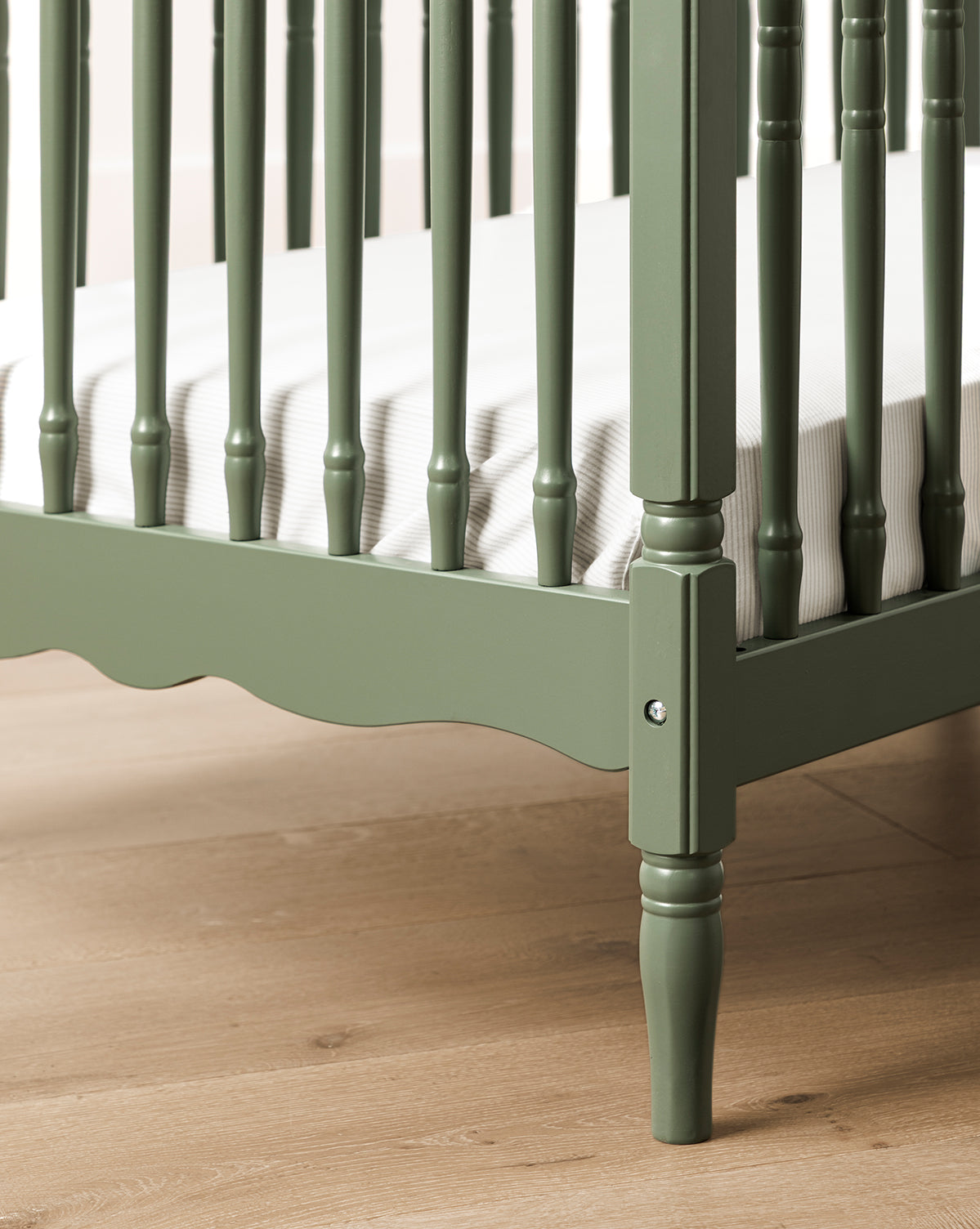 A close-up of the Liberty 3-in-1 Convertible Spindle Crib w/Toddler Bed Conversion Kit in green, crafted from solid pine with vertical slats, decorative spindles, a white striped mattress, and set on a light wooden floor, by McGee & Co.