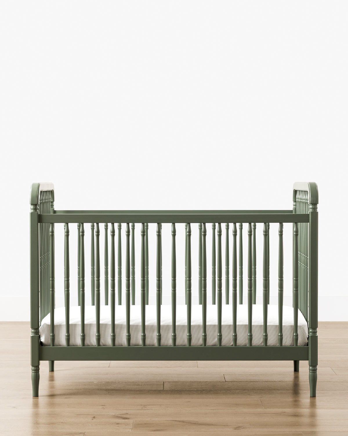The Liberty 3-in-1 Convertible Spindle Crib w/Toddler Bed Conversion Kit in green solid pine, featuring vertical slats and a white mattress, sits on a light wood floor against a plain white wall, by McGee & Co.