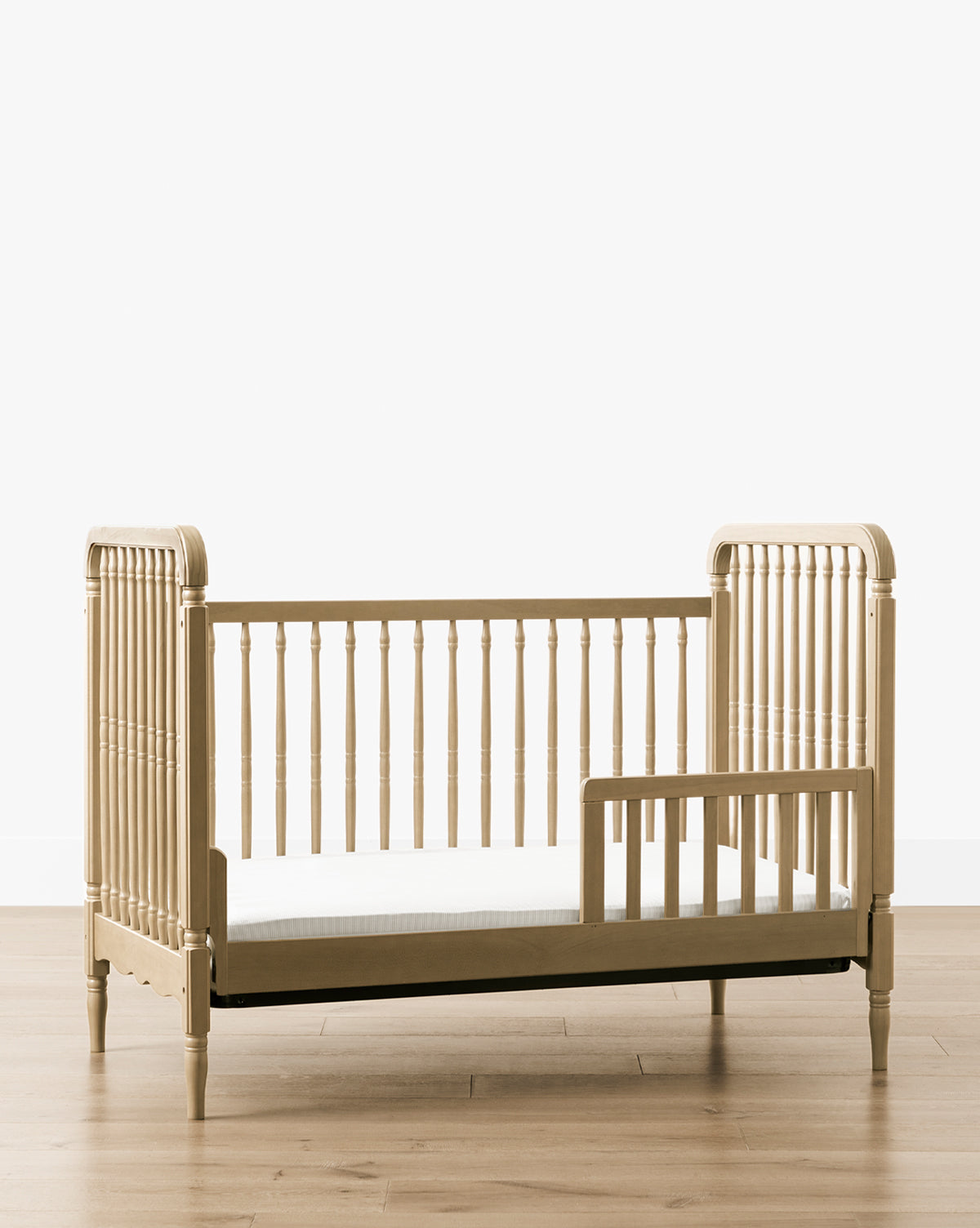 The Liberty 3-in-1 Convertible Spindle Crib w/Toddler Bed Conversion Kit, made from solid pine with vertical slats and a partial safety rail, is shown on a light wood floor against a plain white background by McGee & Co.
