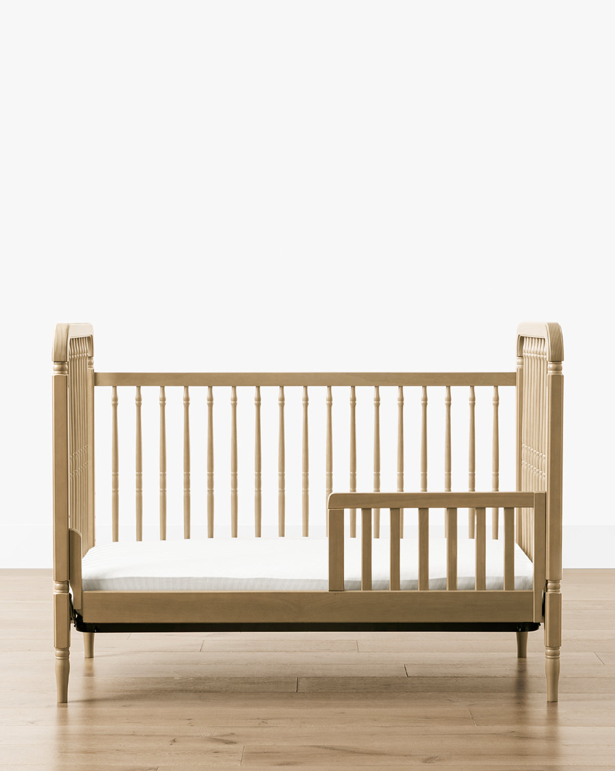 The Liberty 3-in-1 Convertible Spindle Crib with Toddler Bed Conversion Kit, crafted from solid pine, features spindle rails and an open side with a safety guard. It sits on a light wood floor against a plain white background, by McGee & Co.