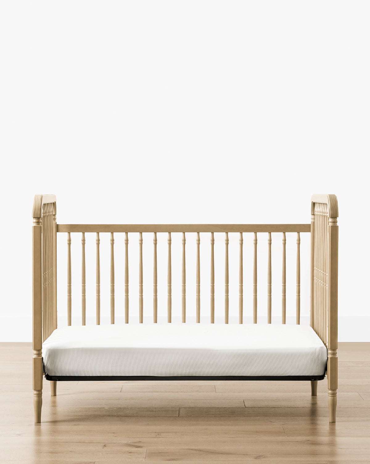 The Liberty 3-in-1 Convertible Spindle Crib w/Toddler Bed Conversion Kit, crafted from solid pine with vertical slats and rounded corners, features a white mattress and is set on a light wood floor against a plain white background, by McGee & Co.