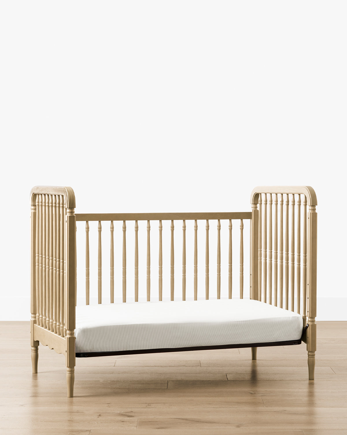 The Liberty 3-in-1 Convertible Spindle Crib w/Toddler Bed Conversion Kit becomes a light solid pine toddler bed with vertical side and back slats, plus a low, open front for easy access. It sits on a wooden floor against a plain white wall, by McGee & Co.