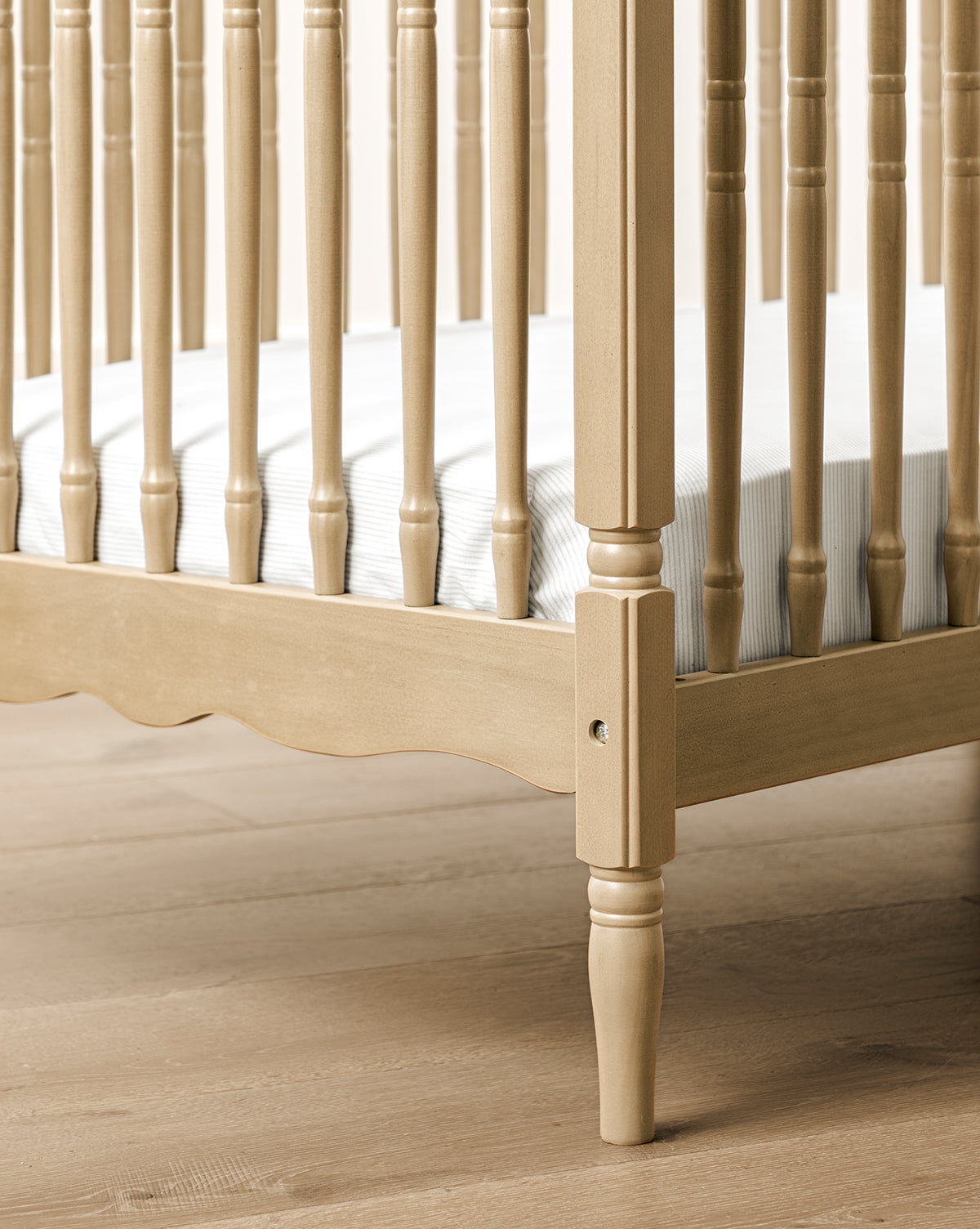 A close-up of the Liberty 3-in-1 Convertible Spindle Crib w/Toddler Bed Conversion Kit, crafted from solid pine with vertical slats, turned legs, and a fitted mattress, shown on a light wood floor by McGee & Co.
