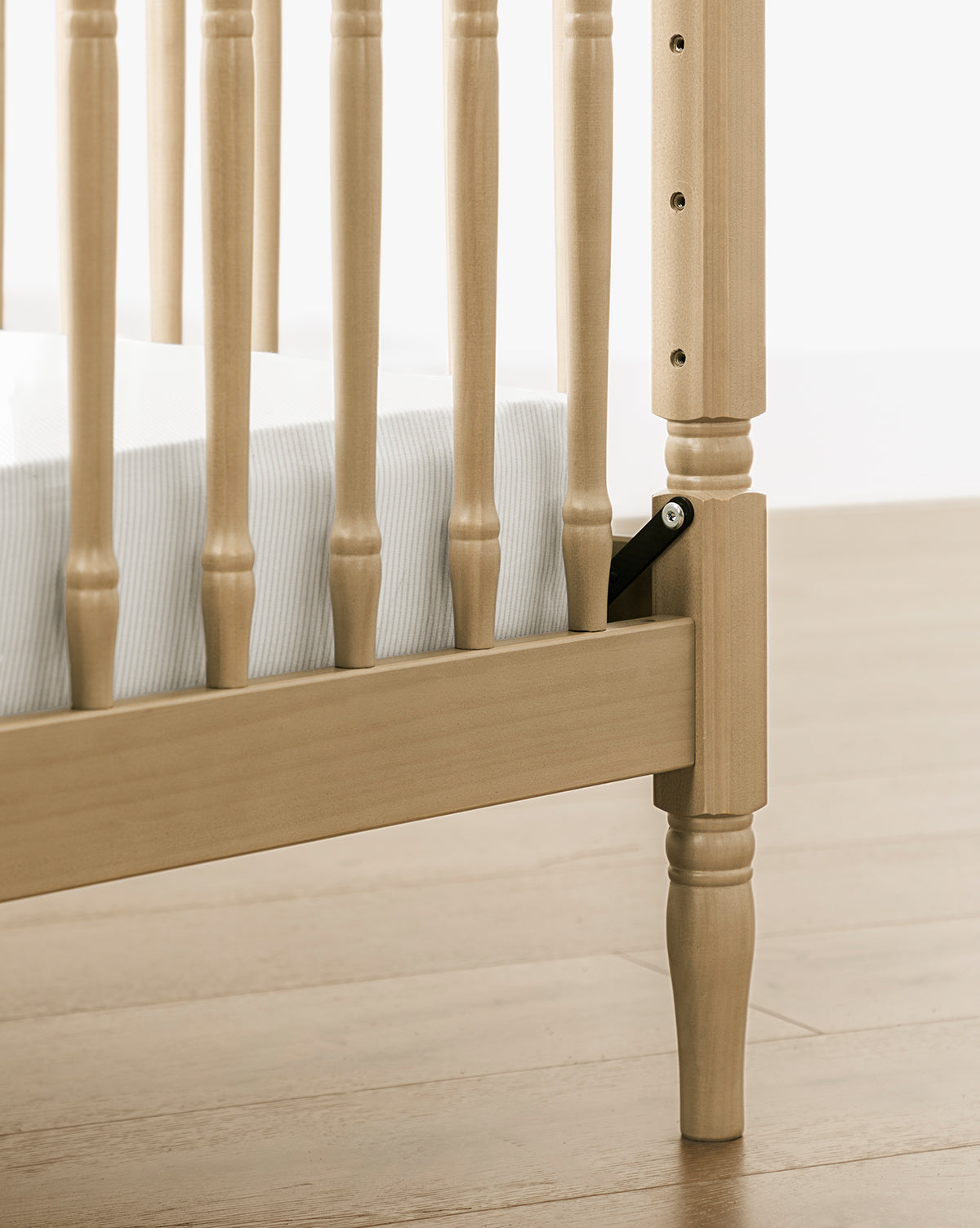Close-up of the Liberty 3-in-1 Convertible Spindle Crib leg and side rail, made from solid pine, with vertical slats. A white mattress sits inside; height adjustment hardware is visible. The crib stands on a light wood floor, by McGee & Co.