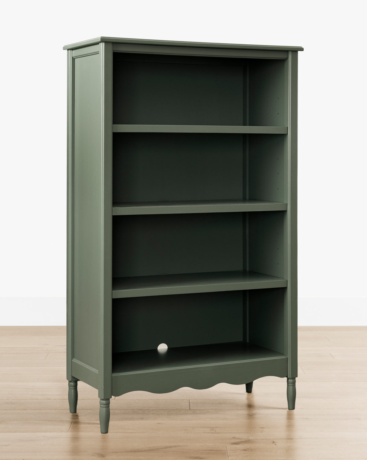 The Liberty Bookcase in green, featuring four shelves, is displayed against a white wall on a light wood floor, highlighting its heirloom craftsmanship. A small white ball rests on the bottom shelf, by McGee & Co.