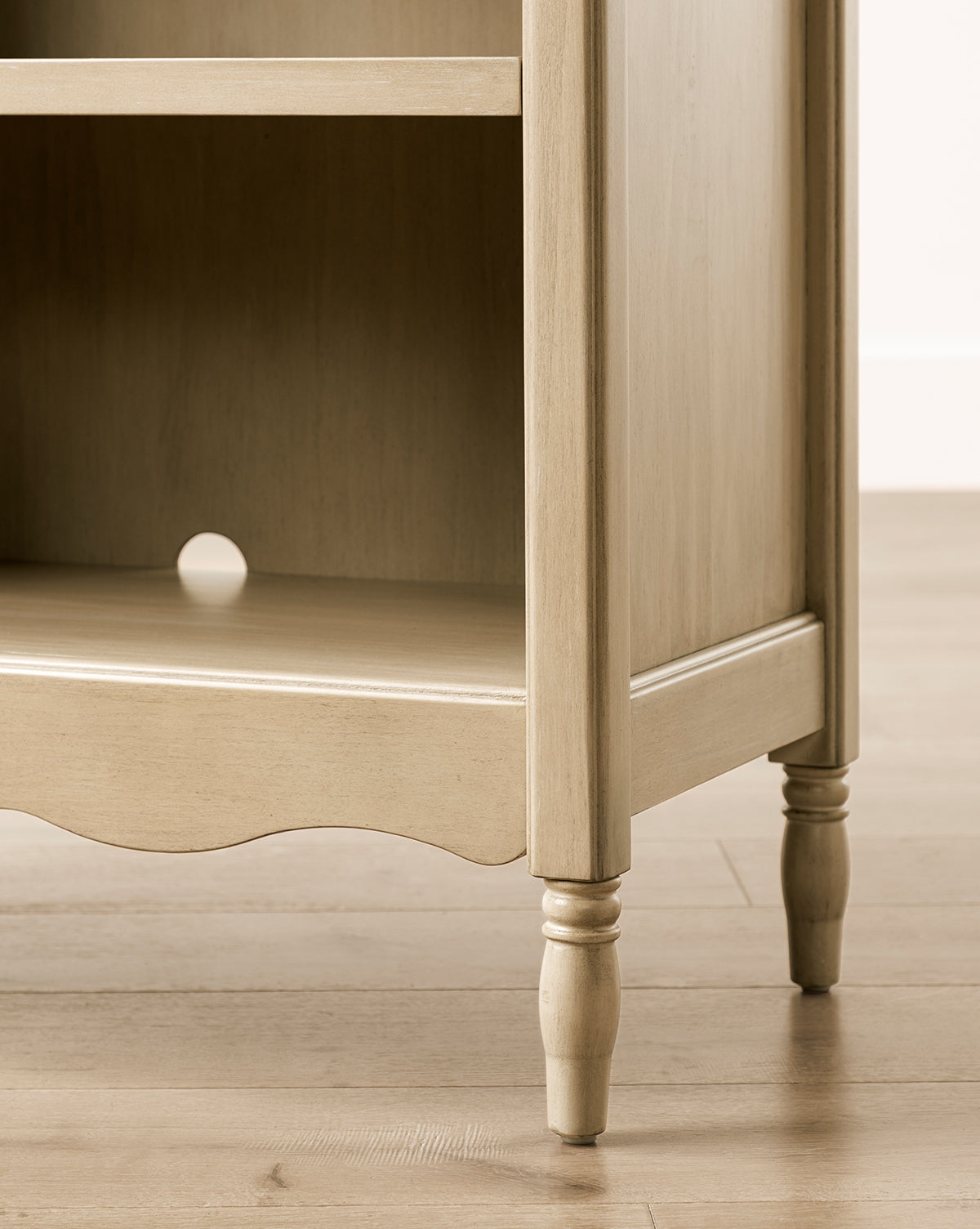 A close-up of the Liberty Bookcase—crafted in light wood with turned legs, an open lower shelf, a scalloped edge, and a rear circular cut-out for cable management—showcasing heirloom-quality craftsmanship on a wooden floor, by McGee & Co.