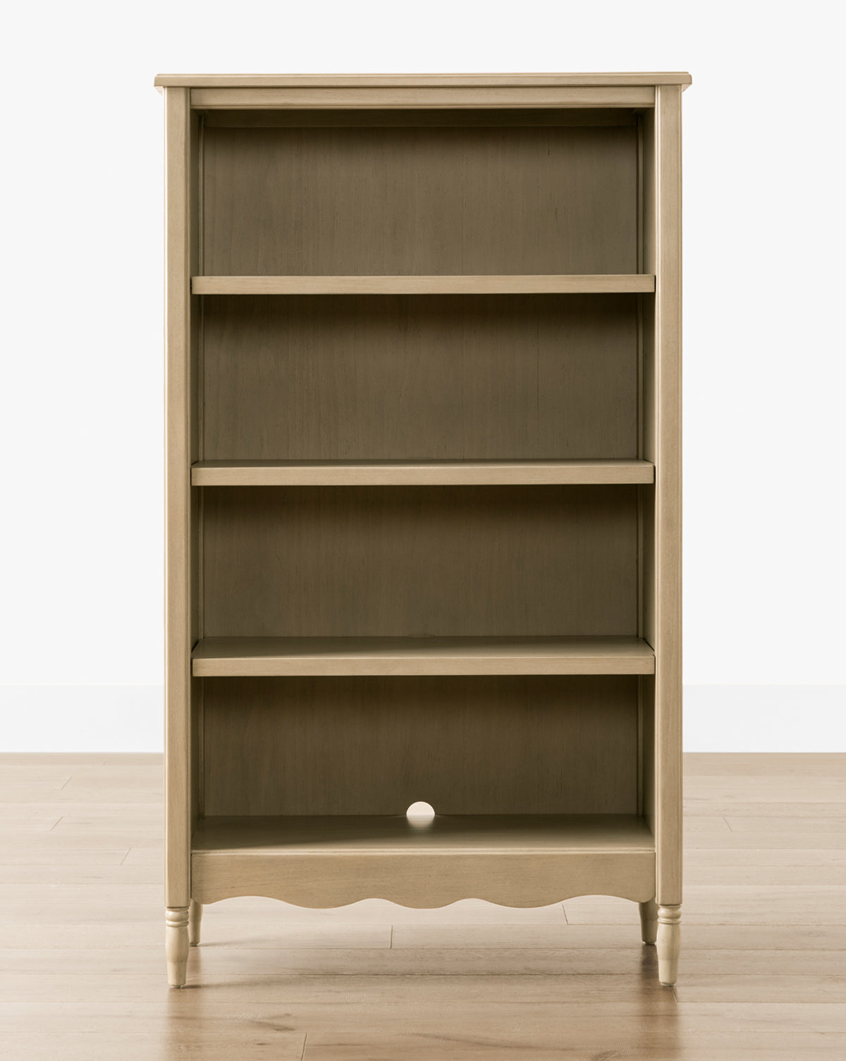The Liberty Bookcase features three shelves and a scalloped bottom edge, set against a wooden floor. Its dark back panel with a small round hole adds distinctive character to this timeless piece, by McGee & Co.