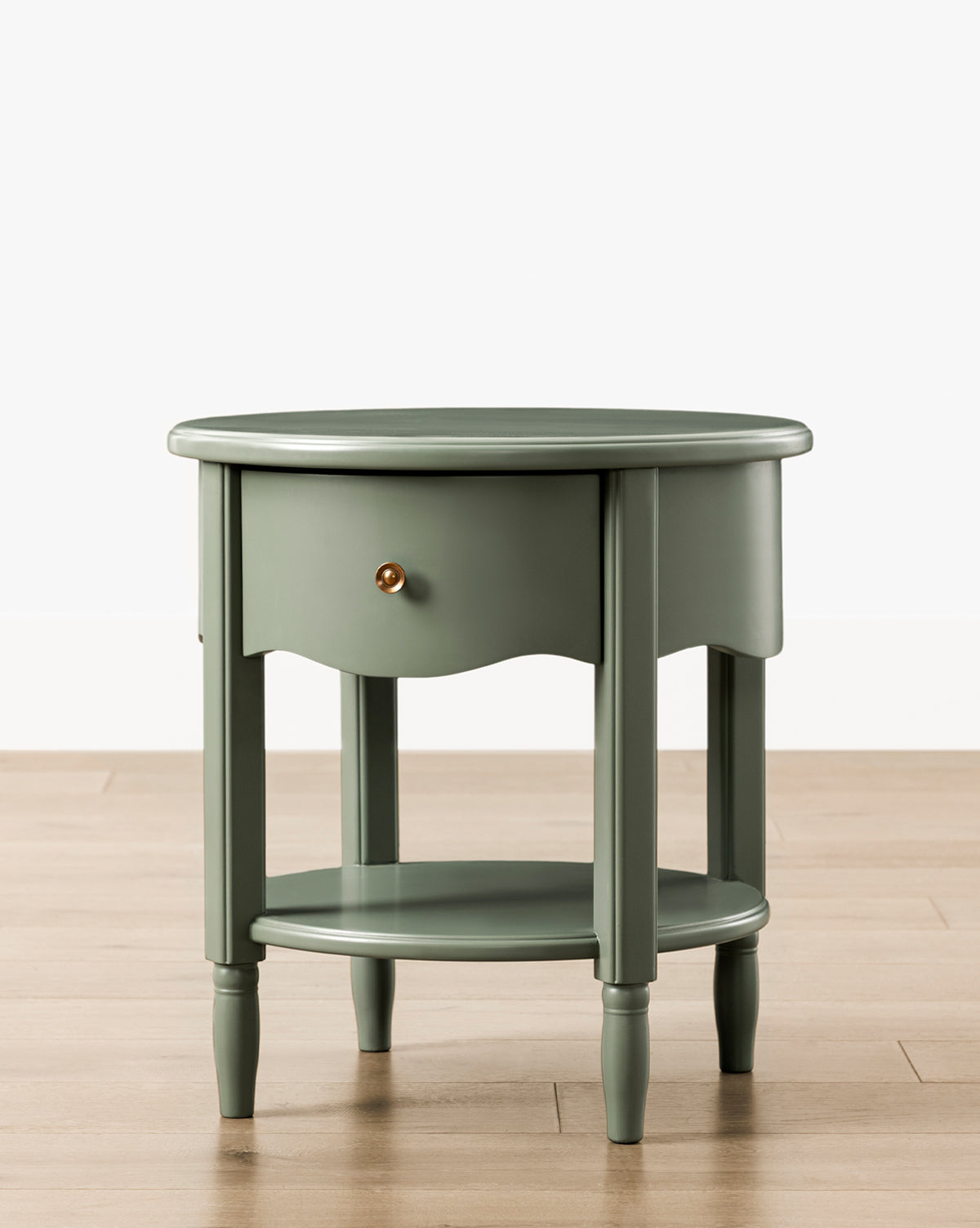 The Liberty Nightstand is a round, green piece with a single drawer and brass knob, lower shelf, and four turned legs. It sits on a light wooden floor against a white wall. GREENGUARD Gold Certified for safer indoor air quality, by McGee & Co.