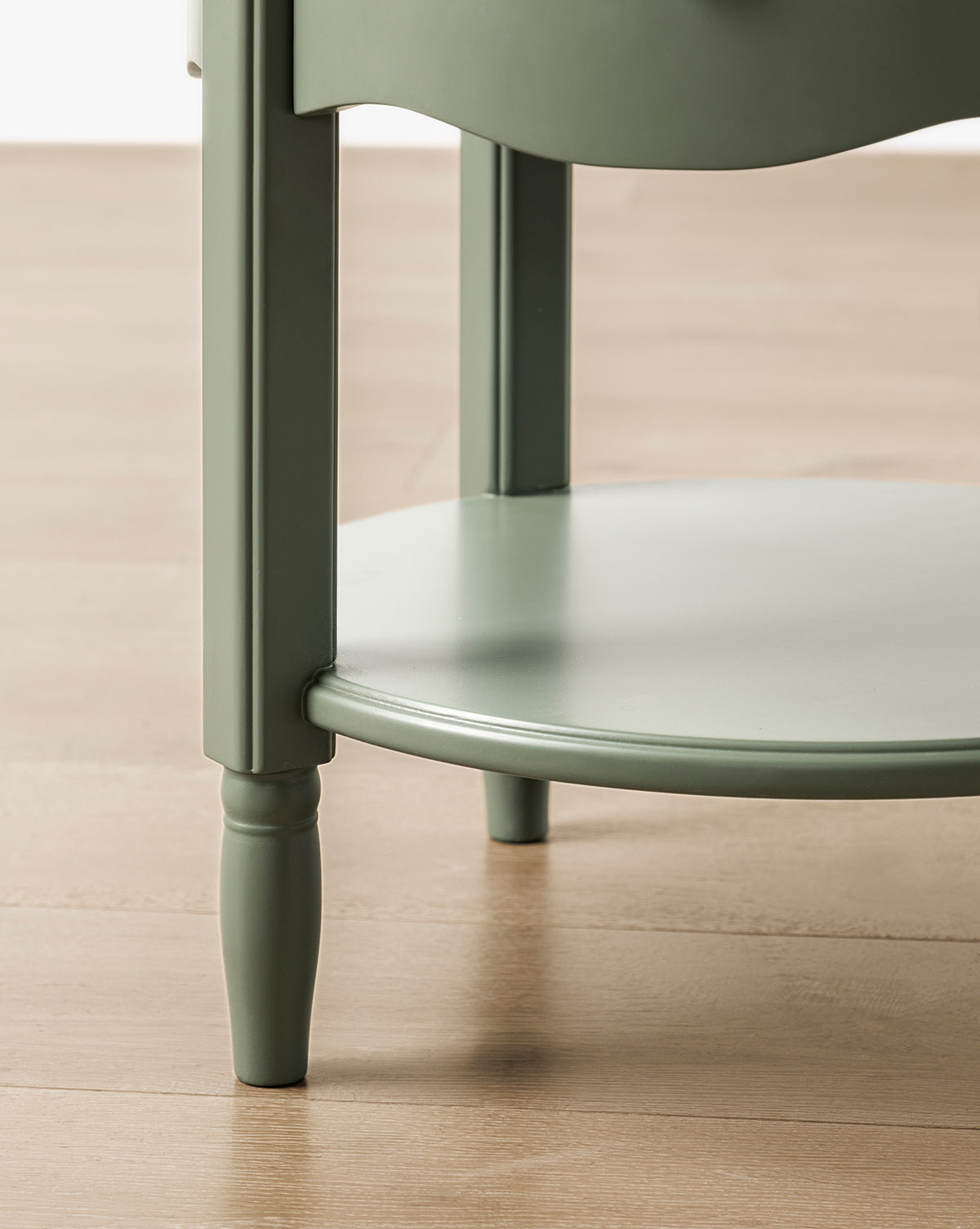 Close-up of the lower section of the GREENGUARD Gold Certified Liberty Nightstand, featuring a green round top, slender legs, and a circular lower shelf—a safe and stylish addition to any nursery on a light wood floor, by McGee & Co.