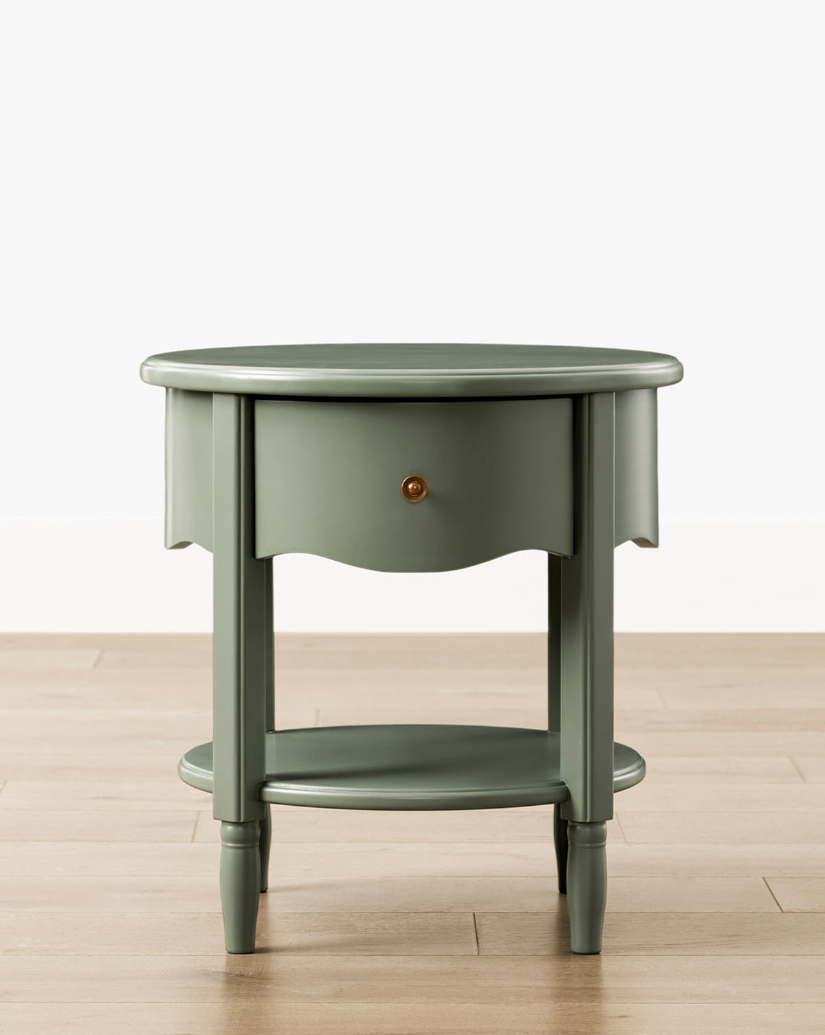The Liberty Nightstand, a round, sage green piece with a small drawer, lower shelf, and four tapered legs, sits on a light wood floor against a white background. GREENGUARD Gold Certified—perfect for nurseries and healthier indoor air by McGee & Co.