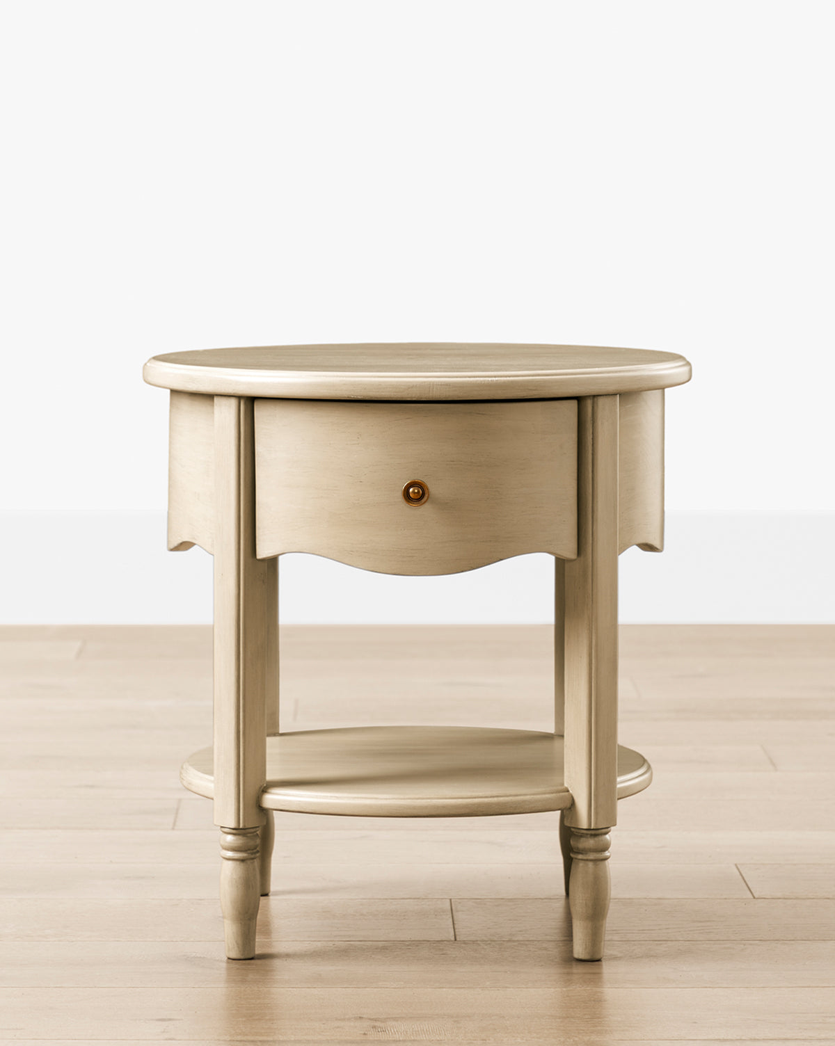 The Liberty Nightstand is a round wooden table with a light, distressed finish, one drawer with a round knob, four legs, and a lower shelf. It sits on a light wood floor against a white background and is GREENGUARD Gold Certified for nurseries by McGee & Co.