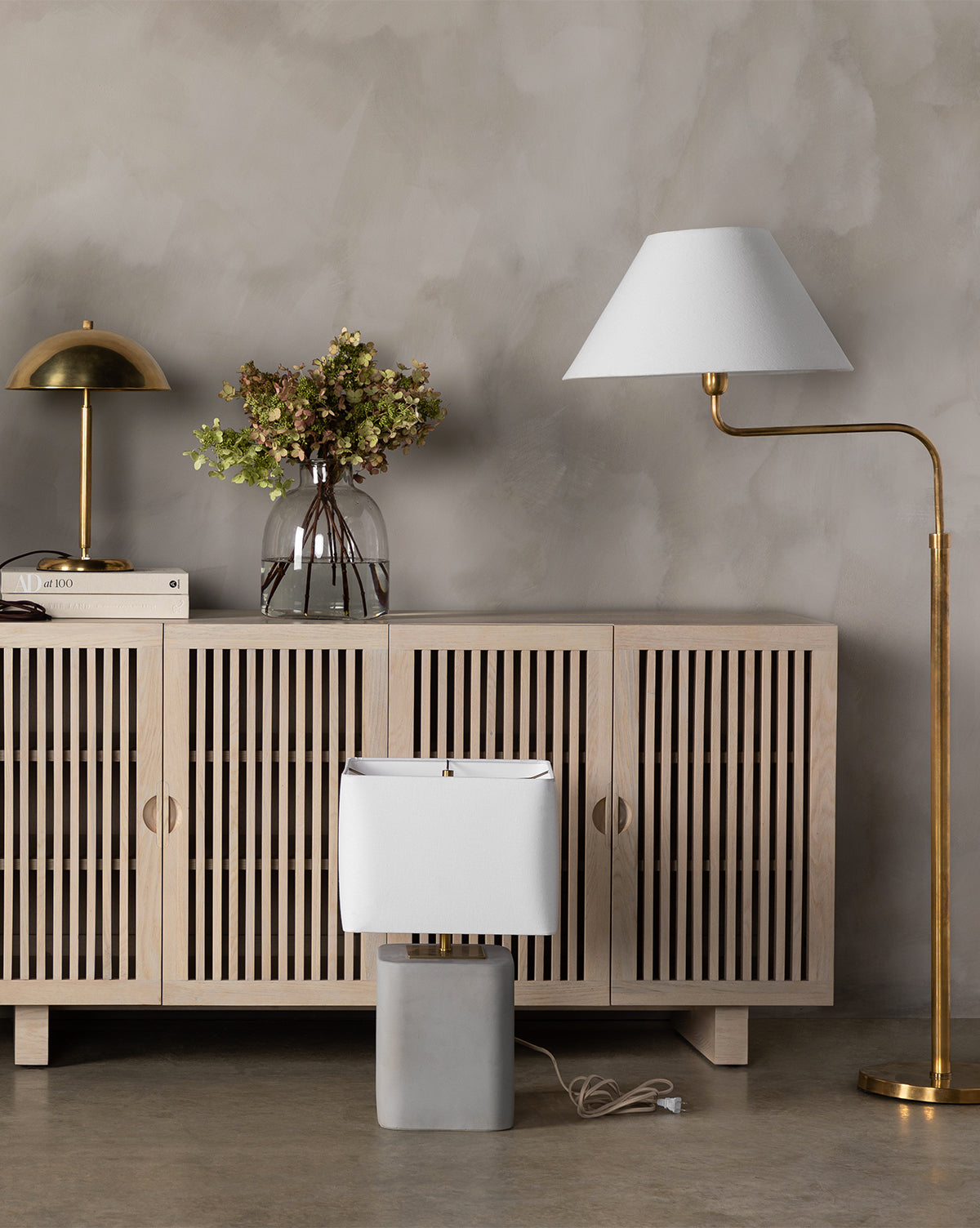 Minimalist interior featuring a light wooden sideboard, brass table lamp, hydrangeas in a glass vase, books, and the Wainwright Swoop Floor Lamp placed on the floor against a gray wall, by McGee & Co.