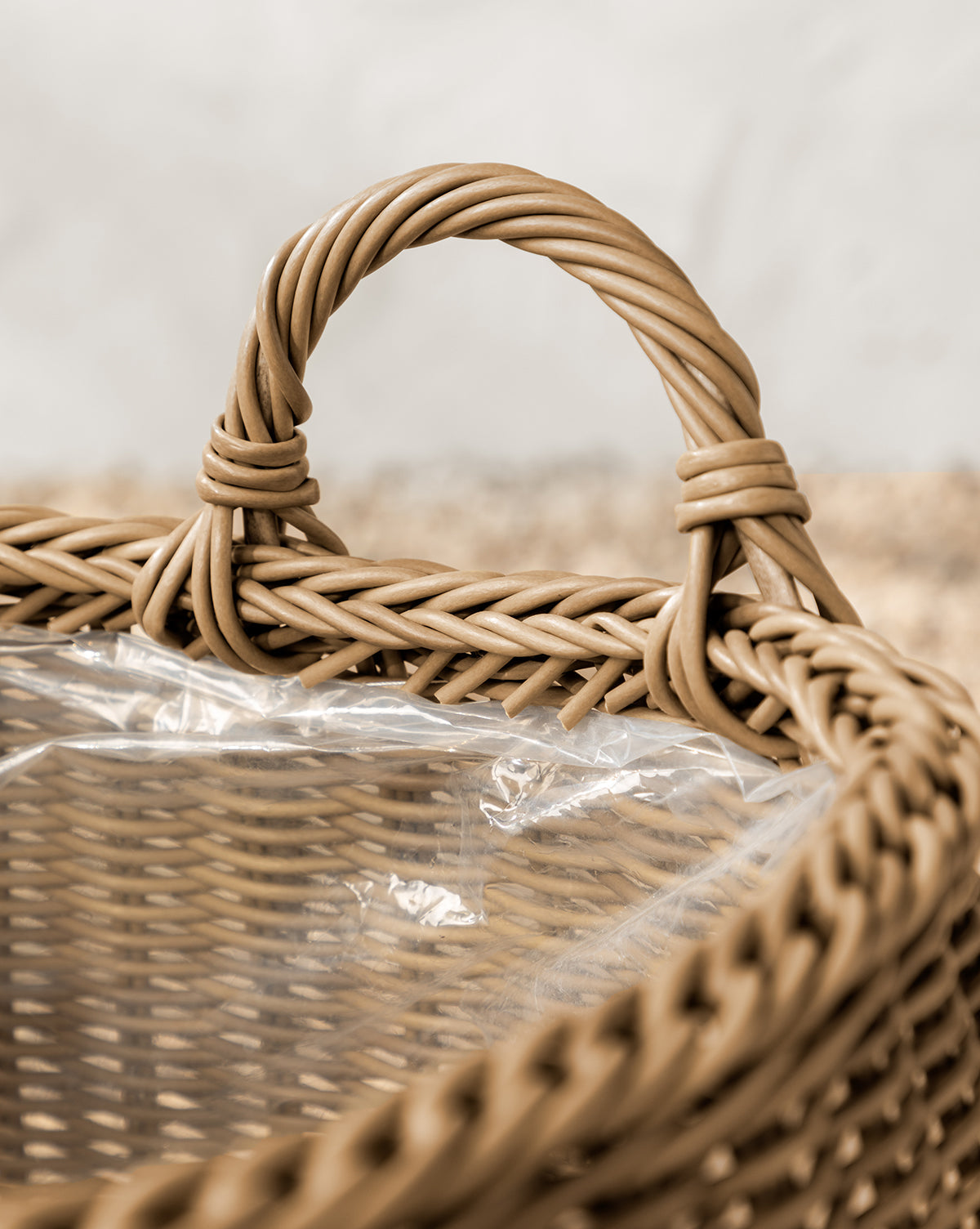 The Lightwash Handled Planter Basket features a sturdy twisted handle and a clear plastic liner, making it perfect for indoor plants. Its poly-rattan texture stands out against a softly blurred, neutral background, by McGee & Co.