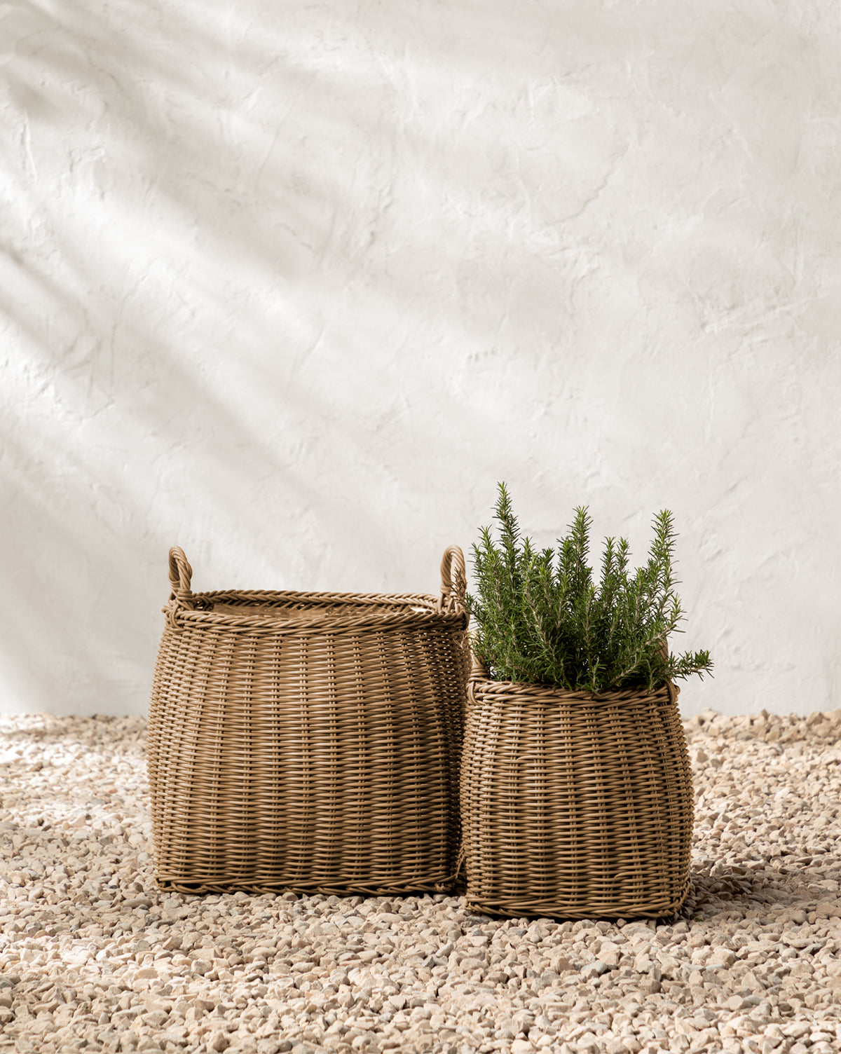 Two Makerspalm Lightwash Handled Planter Baskets rest on pebbled ground; the larger is empty with handles, and the smaller holds a green rosemary plant. Both sit by a textured white wall in soft natural light, McGee & Co.