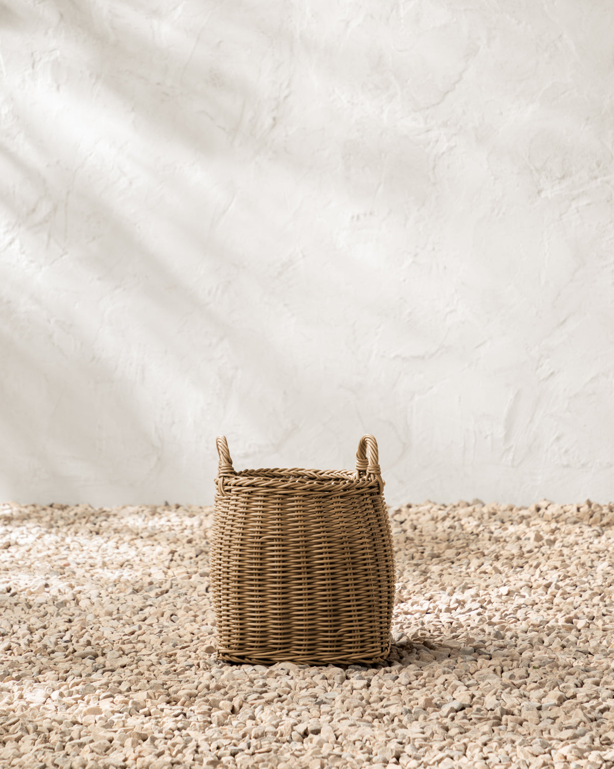 The Lightwash Handled Planter Basket sits on a bed of small pebbles, set against a textured white wall with soft shadows by McGee & Co.