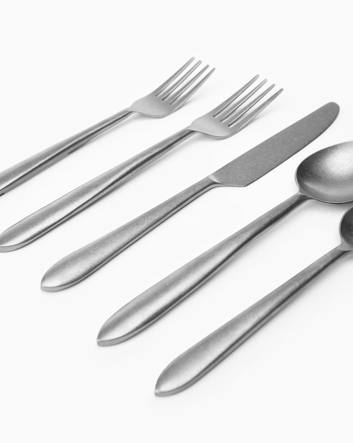 A close-up of the Lilia Flatware (Set of 5)—two forks, a knife, and two spoons—arranged diagonally on a white background.