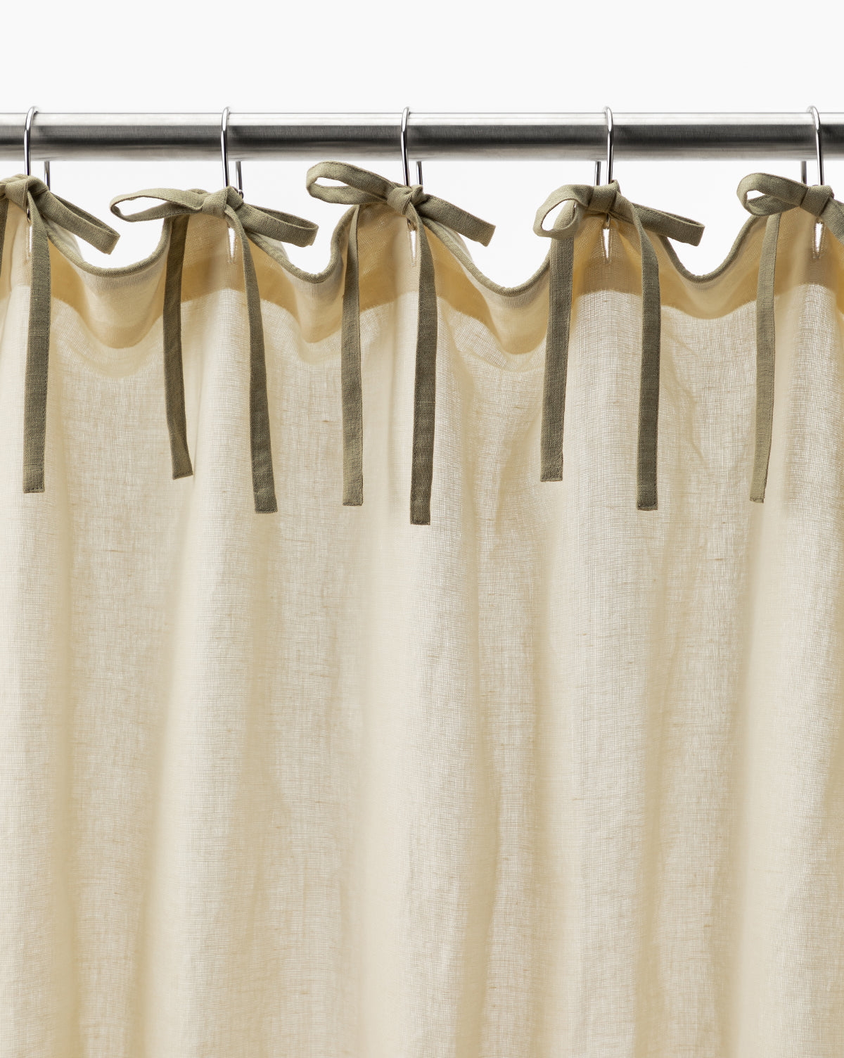 The Lillian Linen Shower Curtain in cream hangs from a metal rod, secured with multiple olive green fabric ties knotted into bows.