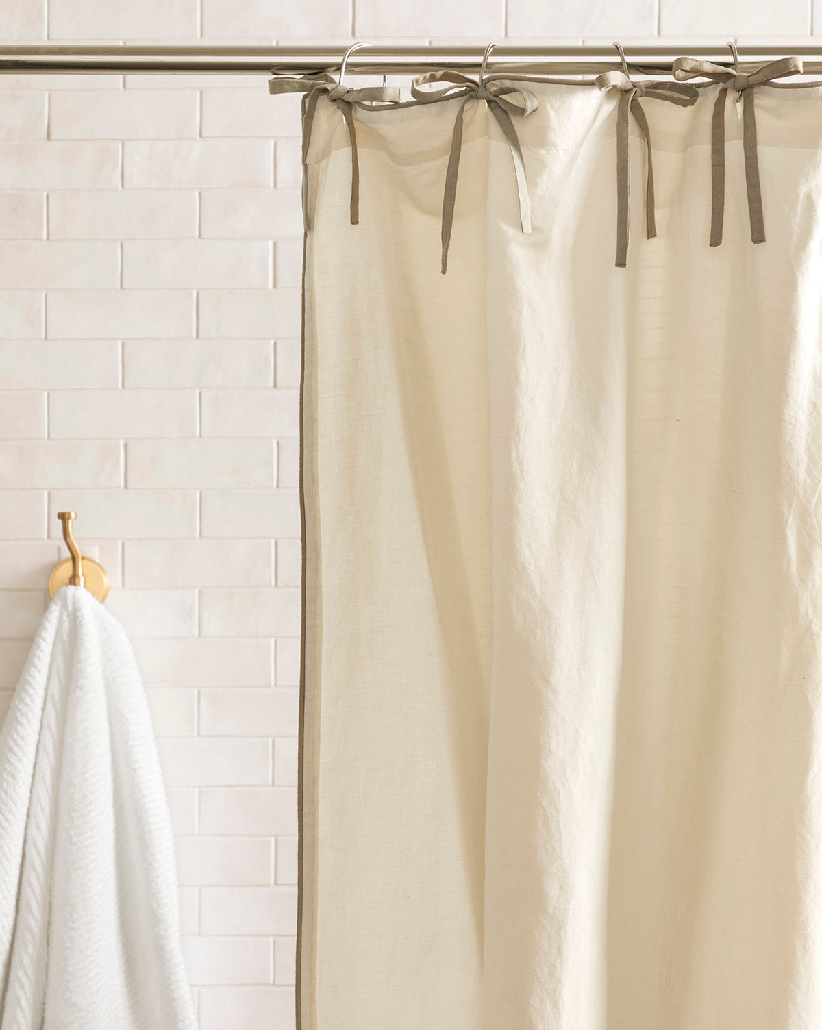 A cream-colored Lillian Linen Shower Curtain with ribbon ties hangs from a silver rod in front of white subway tile, while a white towel rests on a wooden hook nearby.
